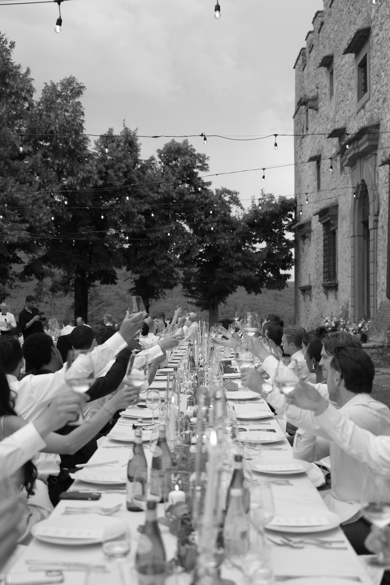 Black and white photo of wedding guests raising glasses in a toast at a long outdoor dining table.