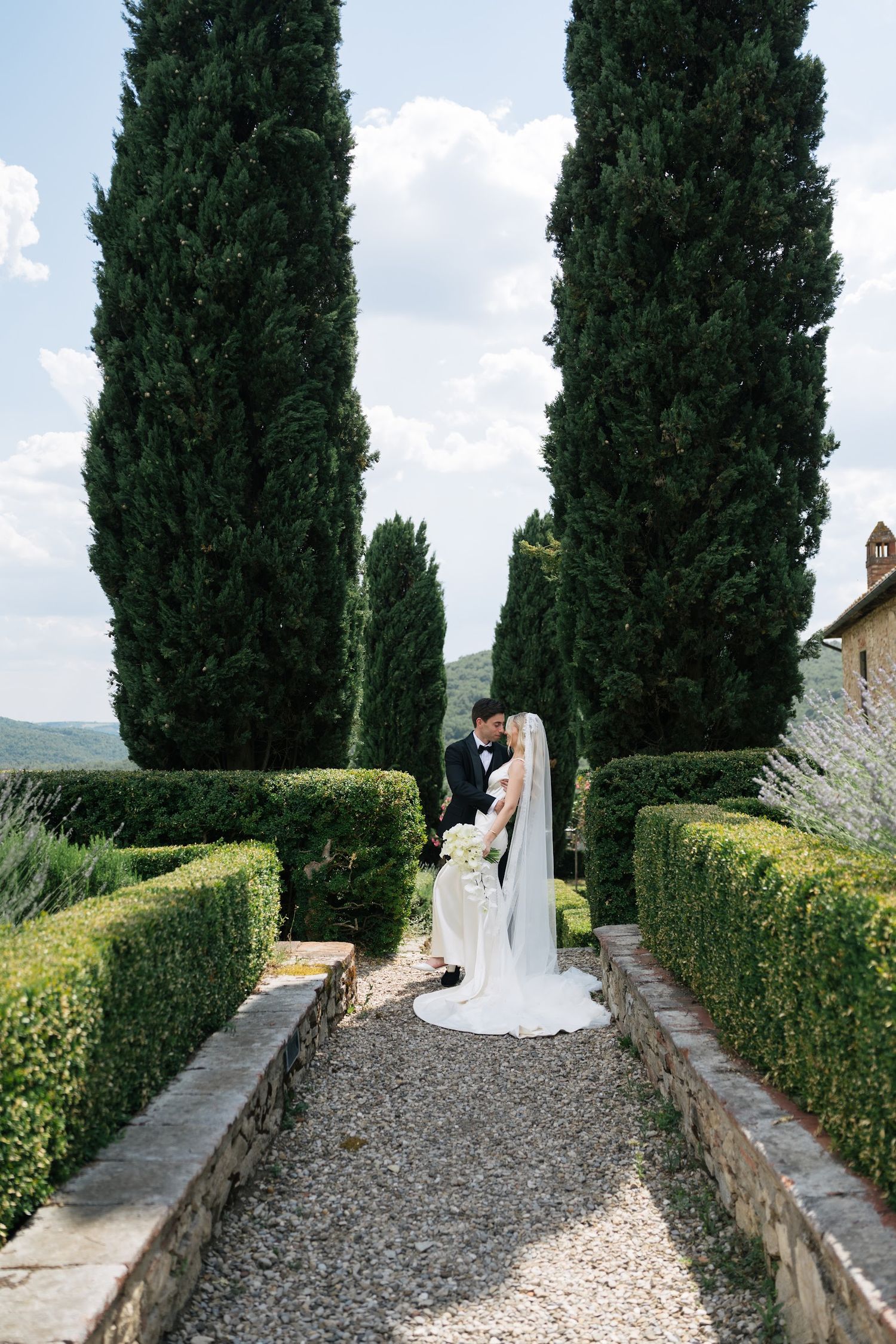 Bride and groom share a romantic moment in a formal Italian garden with tall cypress trees and manicured hedges.