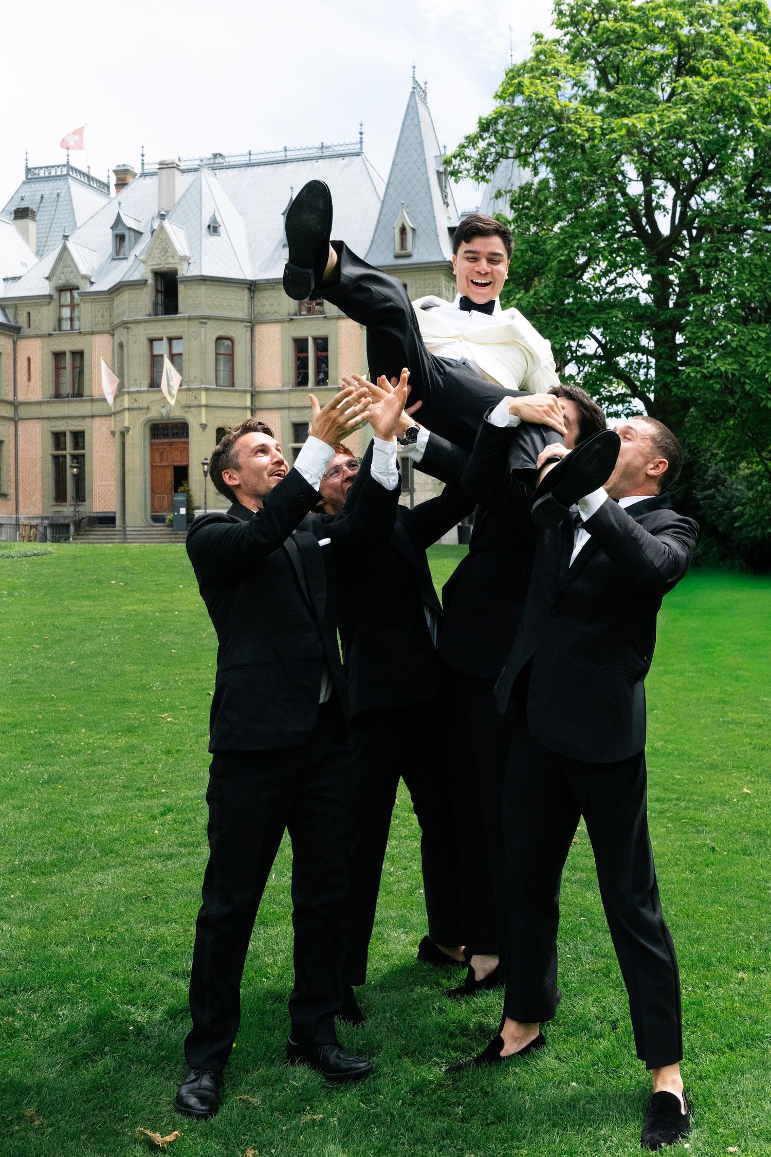 Groomsmen joyfully lifting groom into air outside elegant castle wedding venue on lawn.