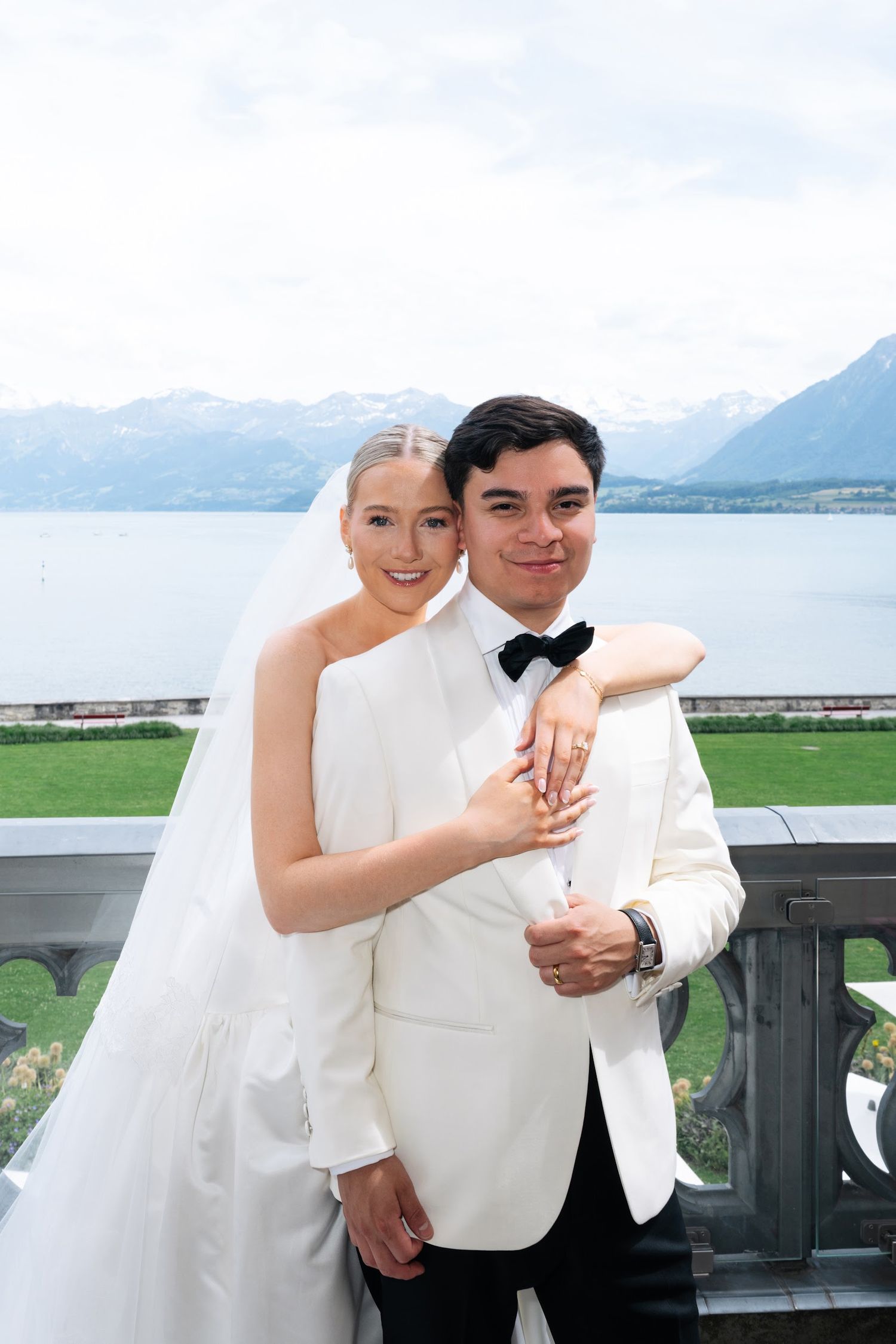 Newlyweds embrace on balcony overlooking alpine lake, groom in white jacket, bride in strapless gown with veil and ring.