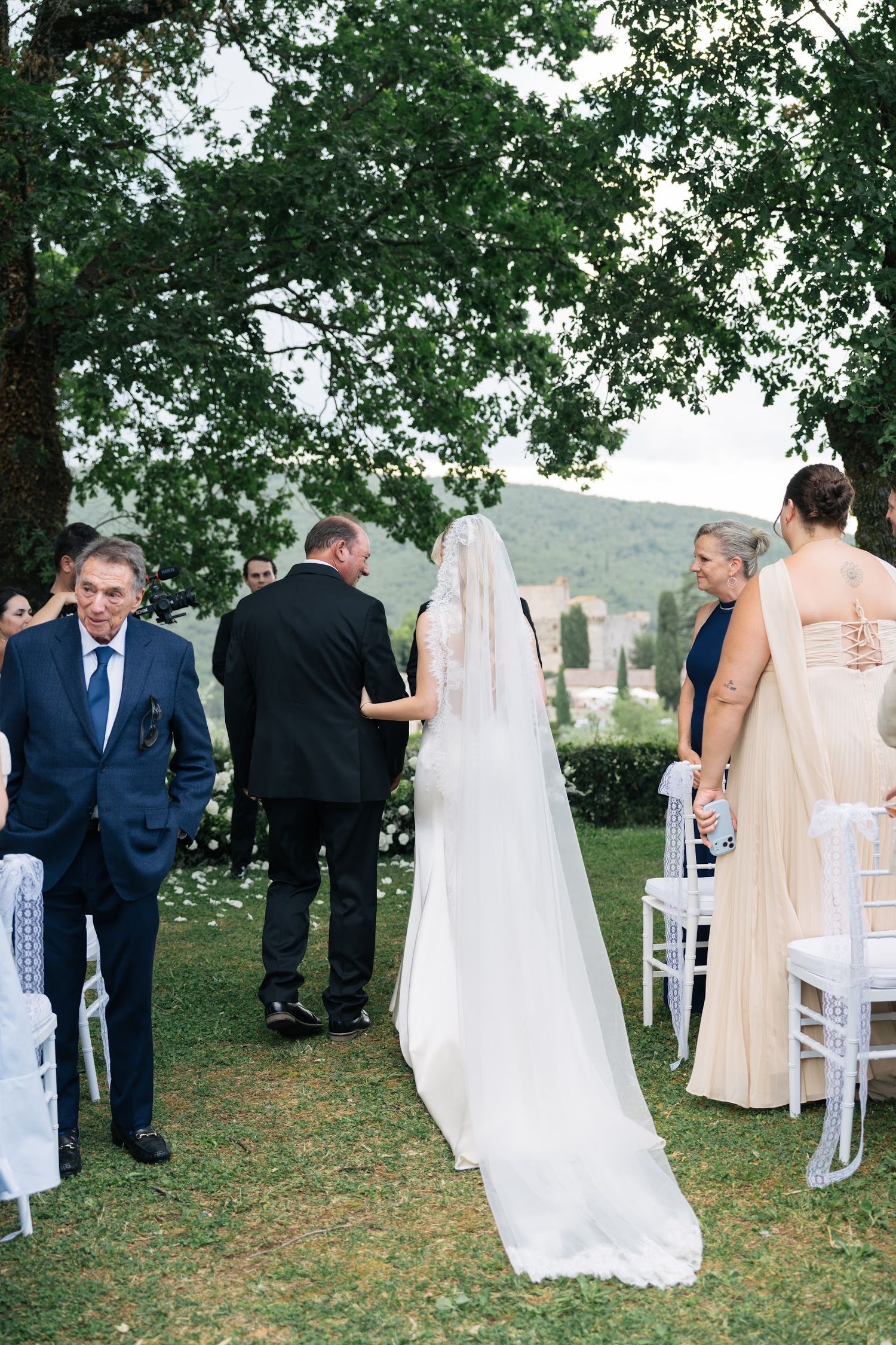 Bride in white gown and veil walks down outdoor aisle escorted by family at a Tuscan countryside wedding.