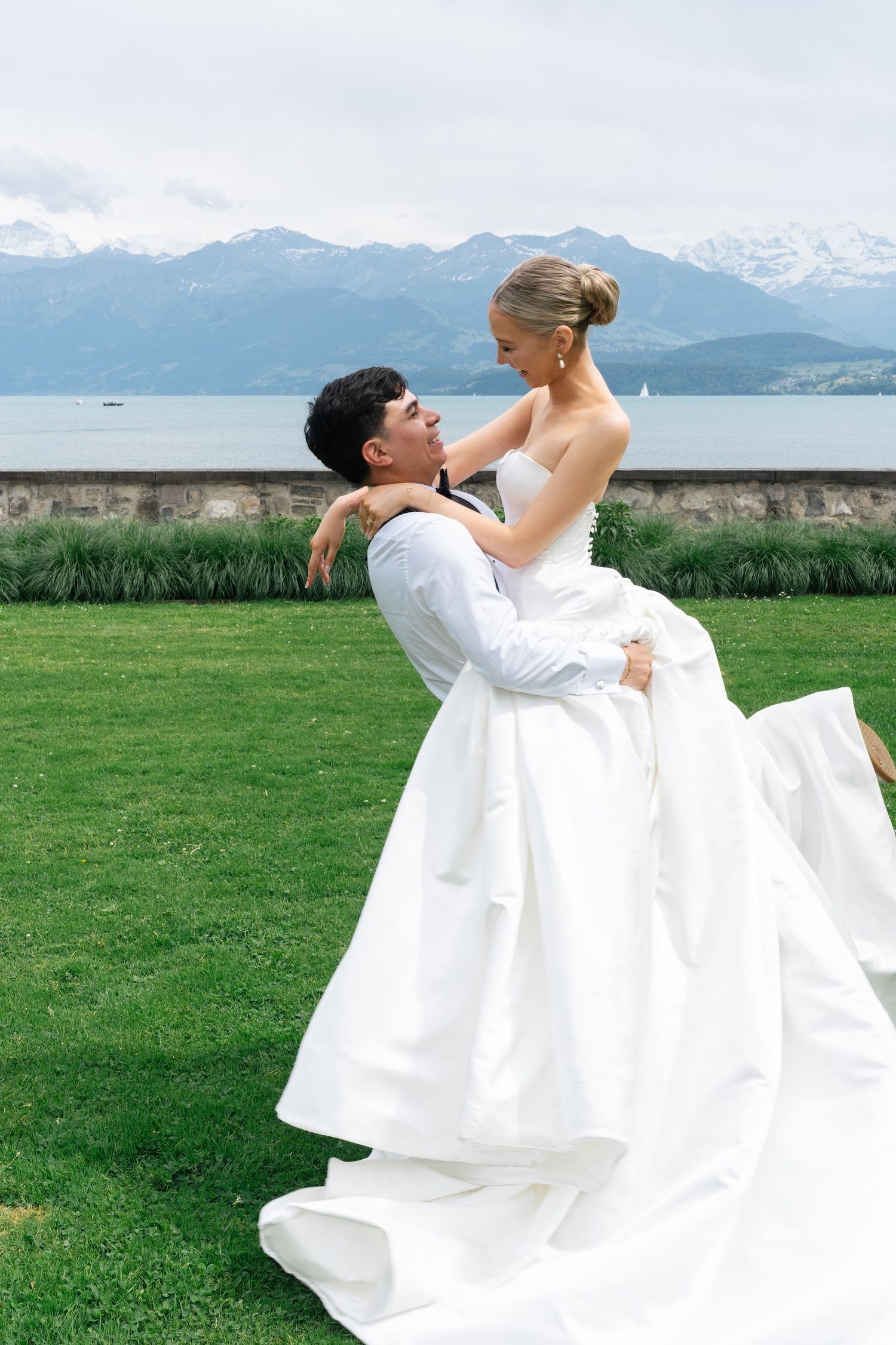 Groom lifting bride in white ballgown on green lawn with alpine lake and snow-capped mountains backdrop.
