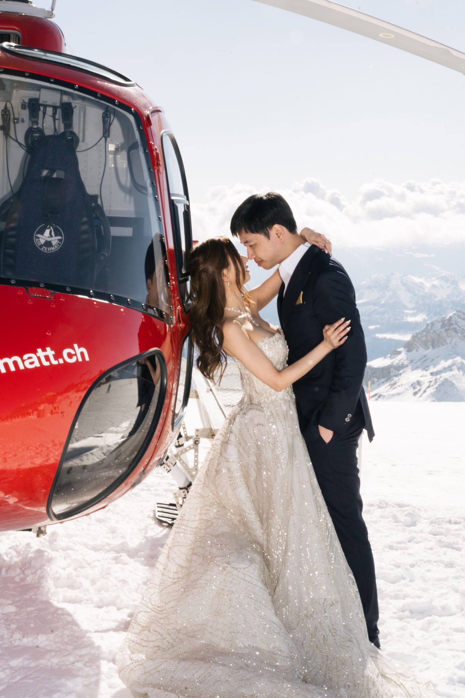 Bride and groom embrace romantically beside red helicopter on snowy alpine mountain summit in Switzerland.