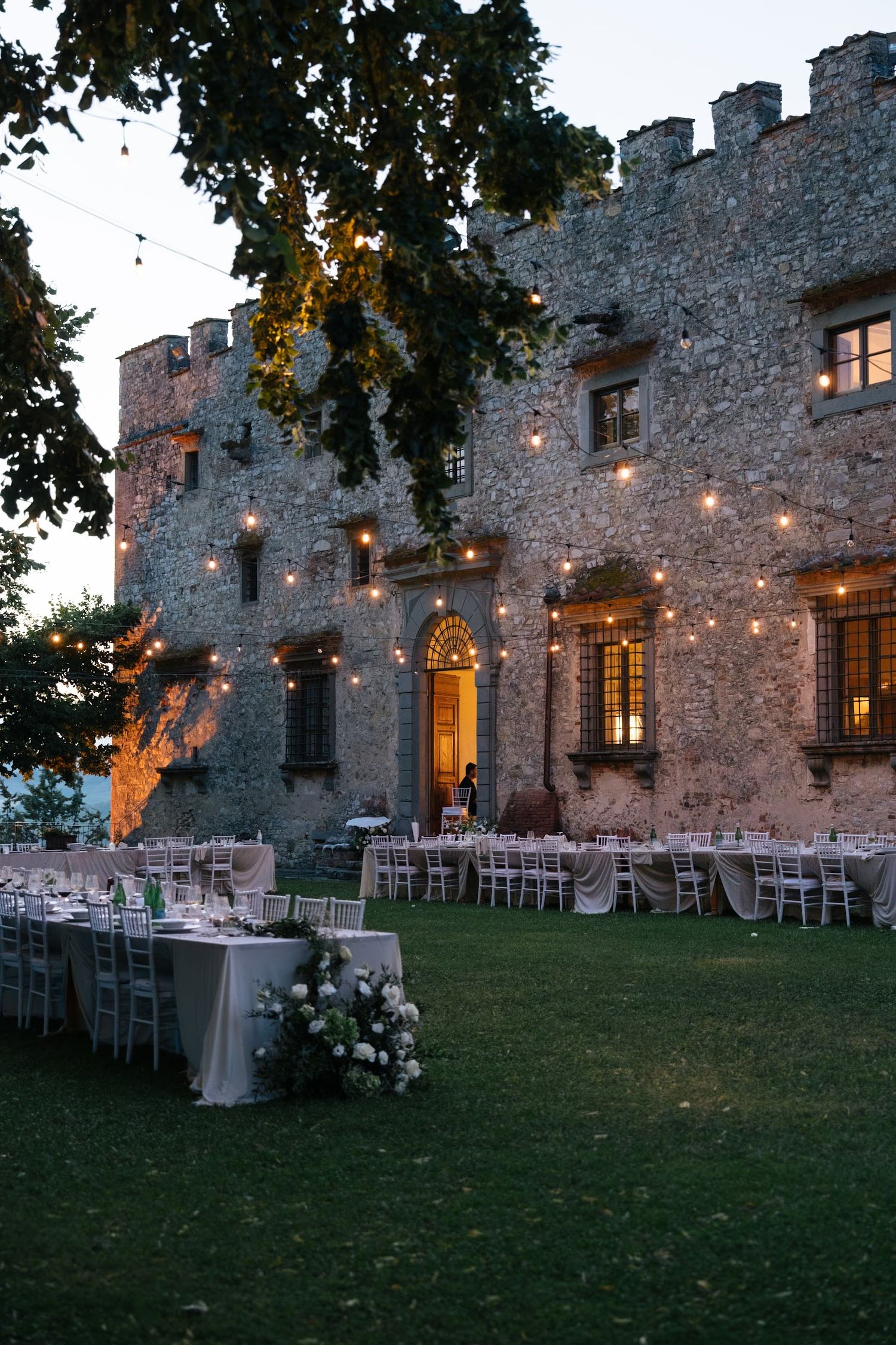 Outdoor wedding reception tables set at dusk beside historic Italian stone castle with string lights.