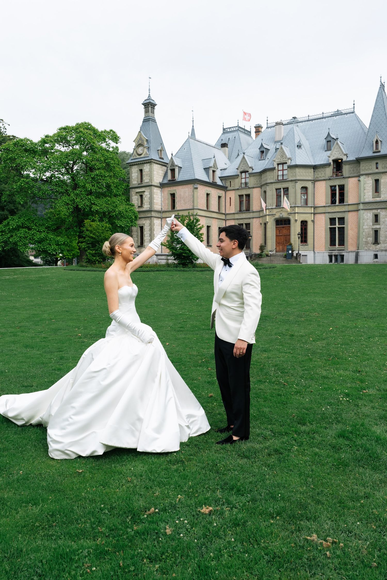 Bride and groom twirling on green lawn in front of elegant castle wedding venue in Switzerland.