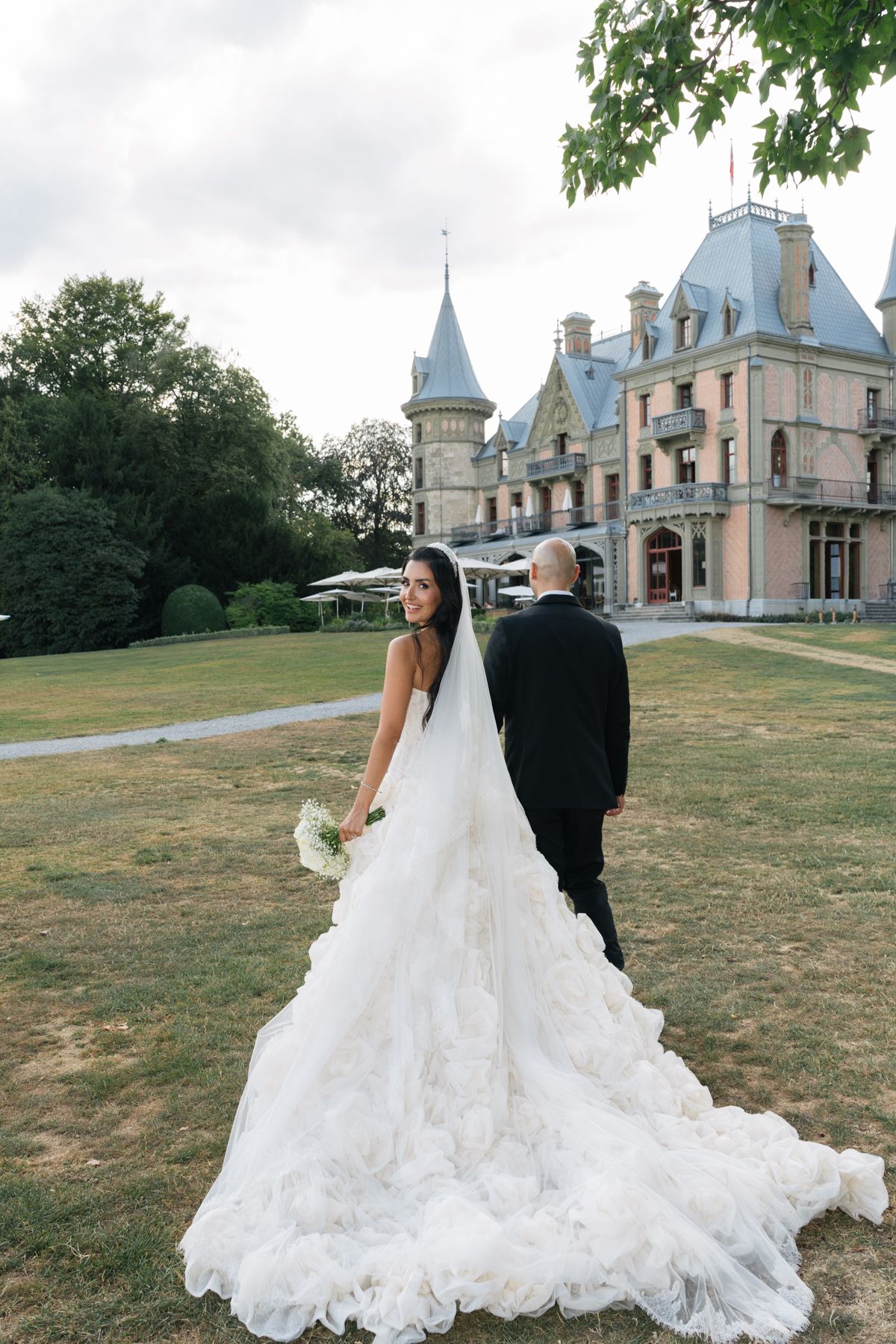 Bride in a flowing ruffled gown and groom walk toward a fairytale castle venue on green grounds.
