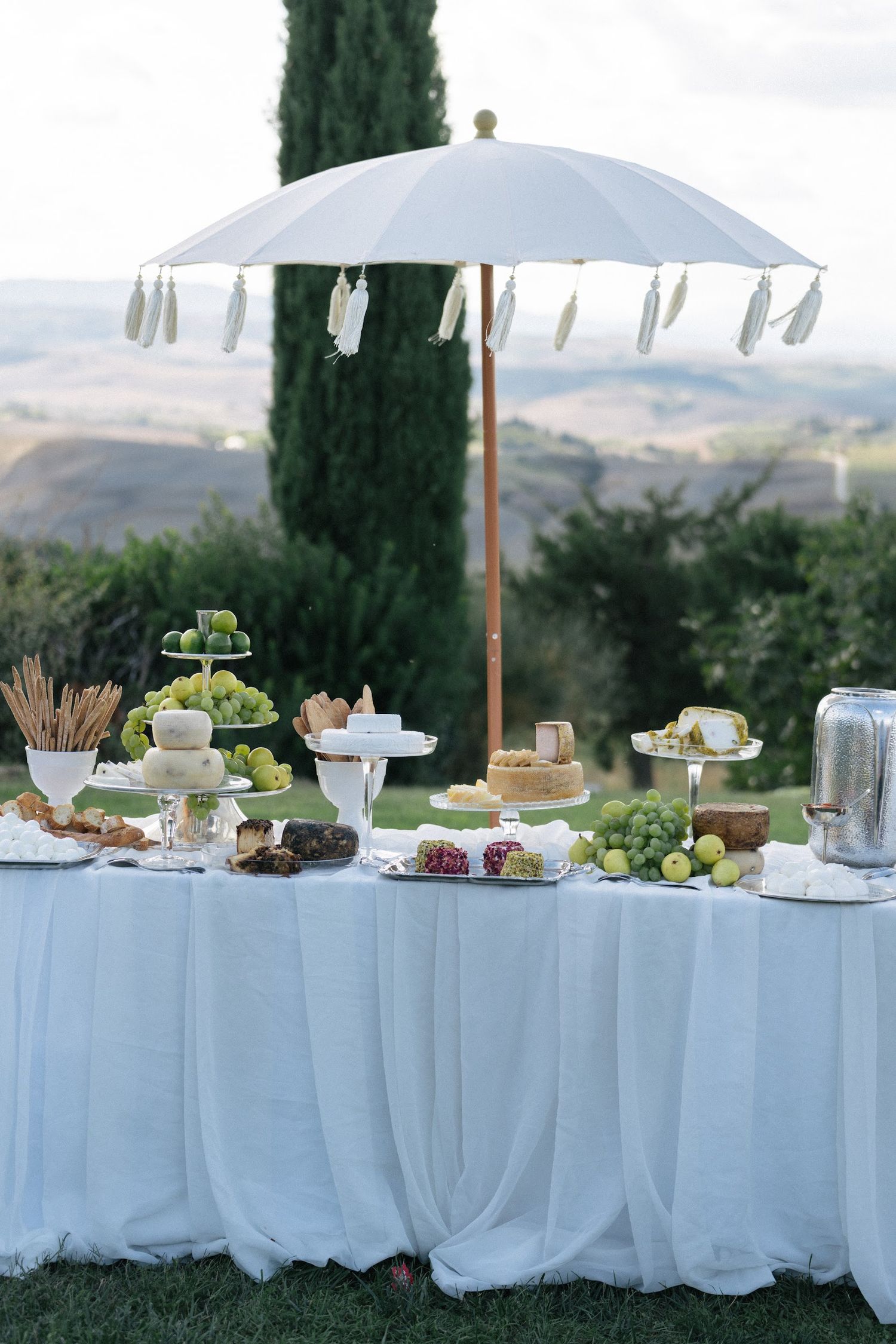 Elegant outdoor wedding grazing table with white umbrella, fruits, and cheeses overlooking Tuscan hills.