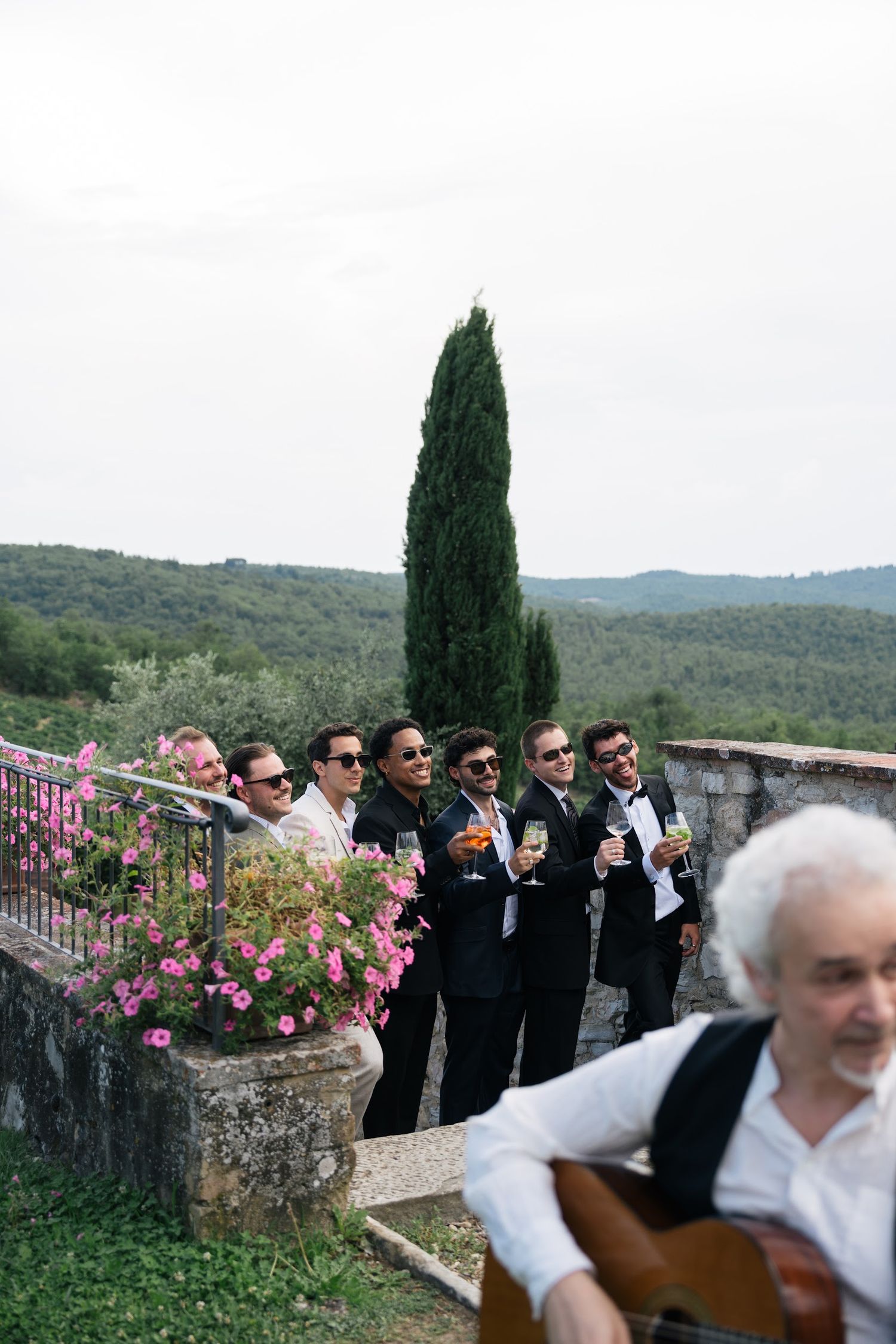 Groomsmen in tuxedos holding drinks on a stone terrace overlooking Tuscan hills with a guitarist.