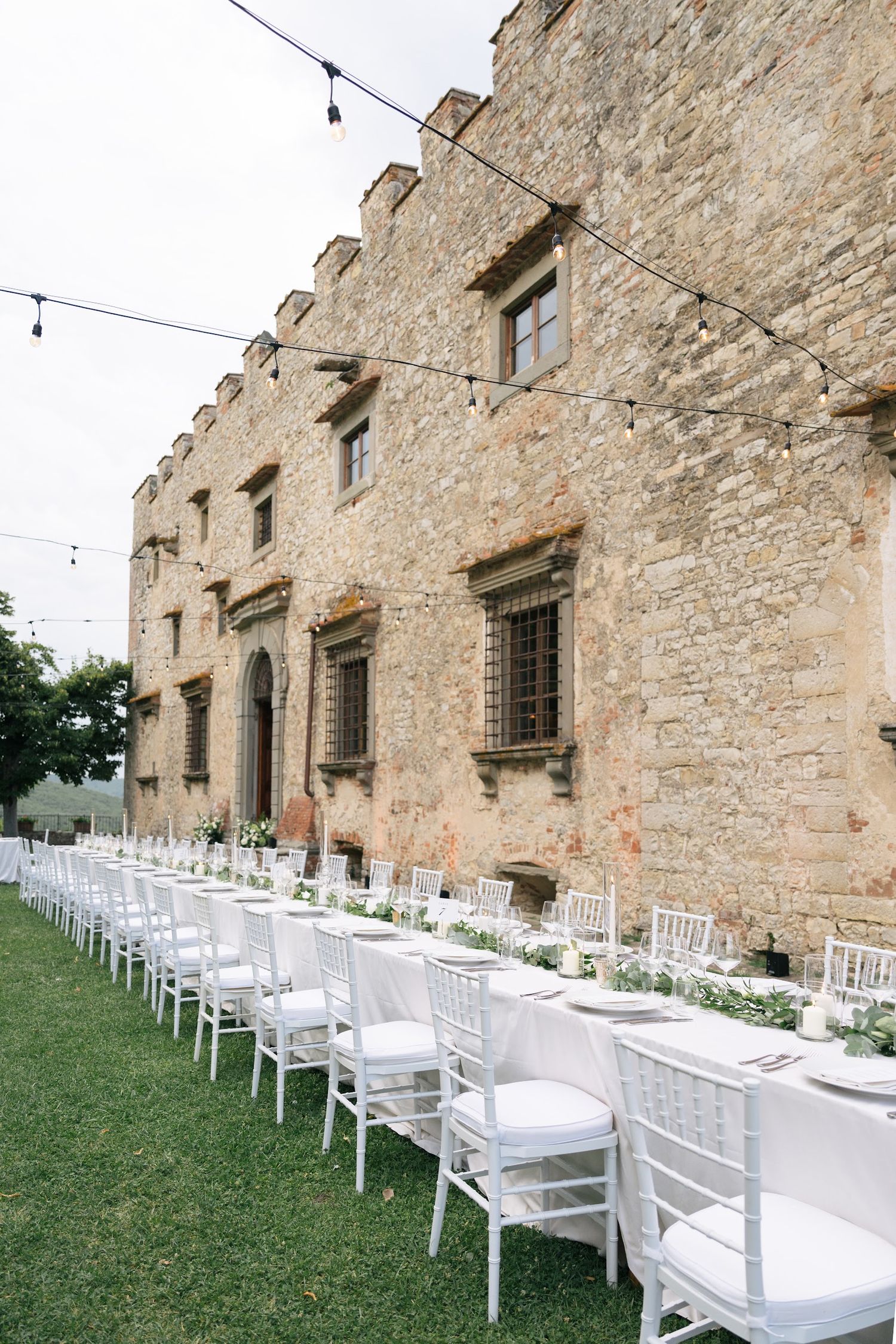 Long outdoor wedding reception tables with white chiavari chairs set against a historic stone villa.