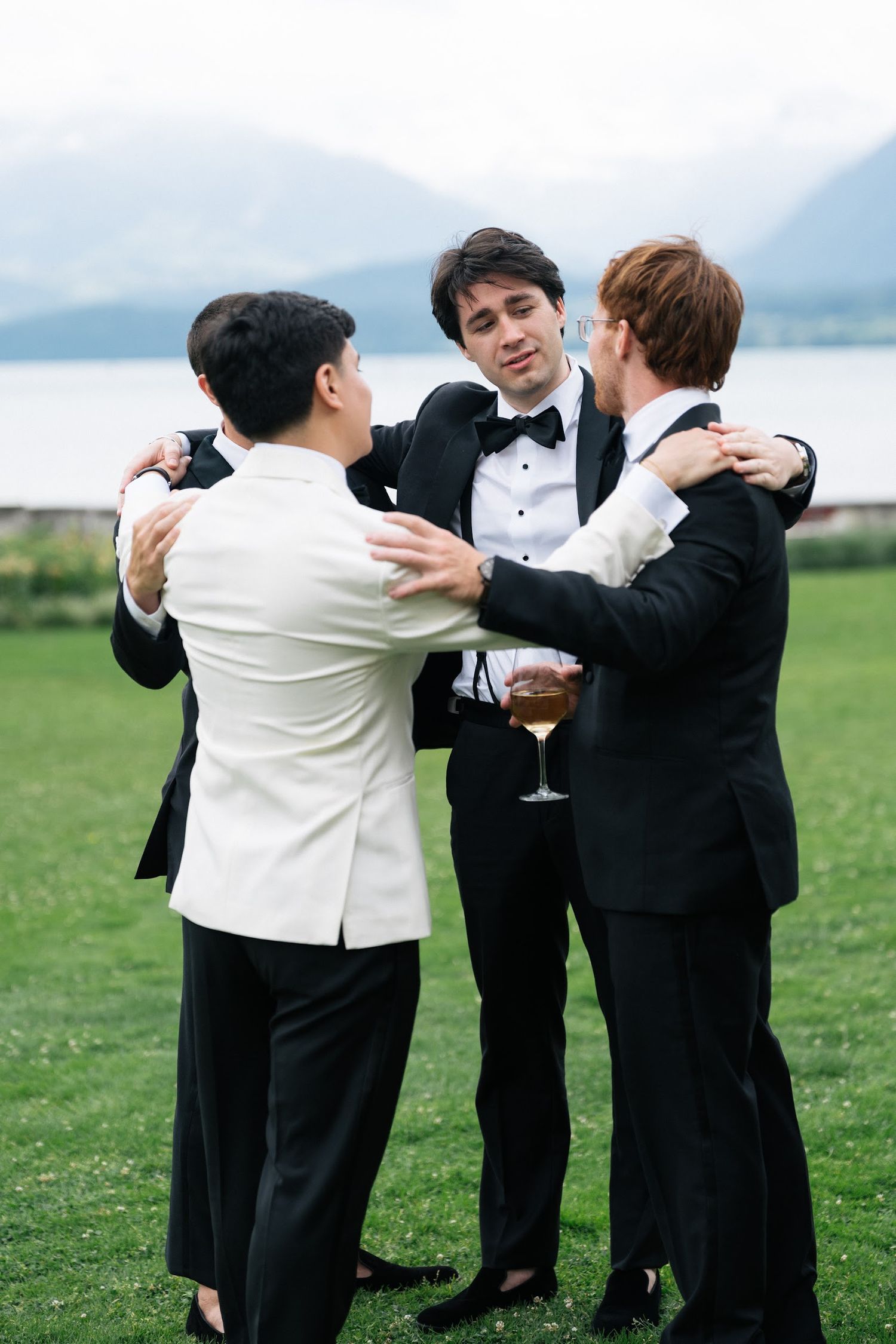 Groom in white jacket embraces two friends in black tuxedos on a green lawn with mountain lake backdrop.