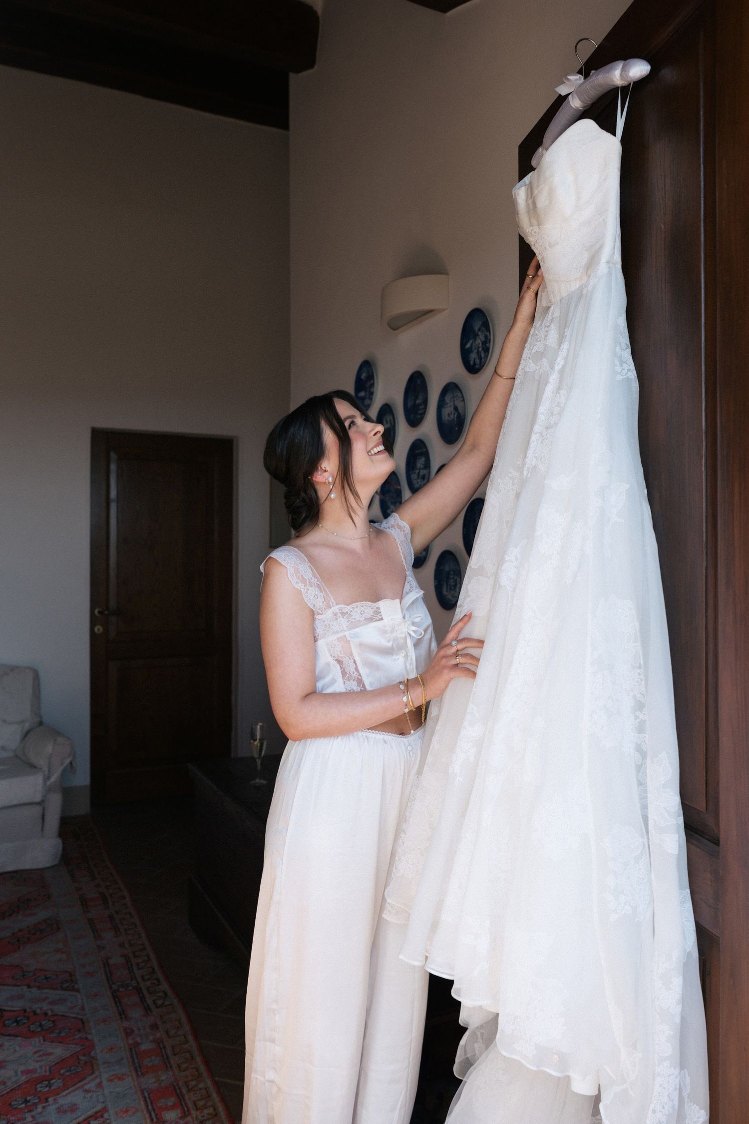 Excited bride in white slip dress reaching up to touch her lace wedding gown hanging in a rustic room.