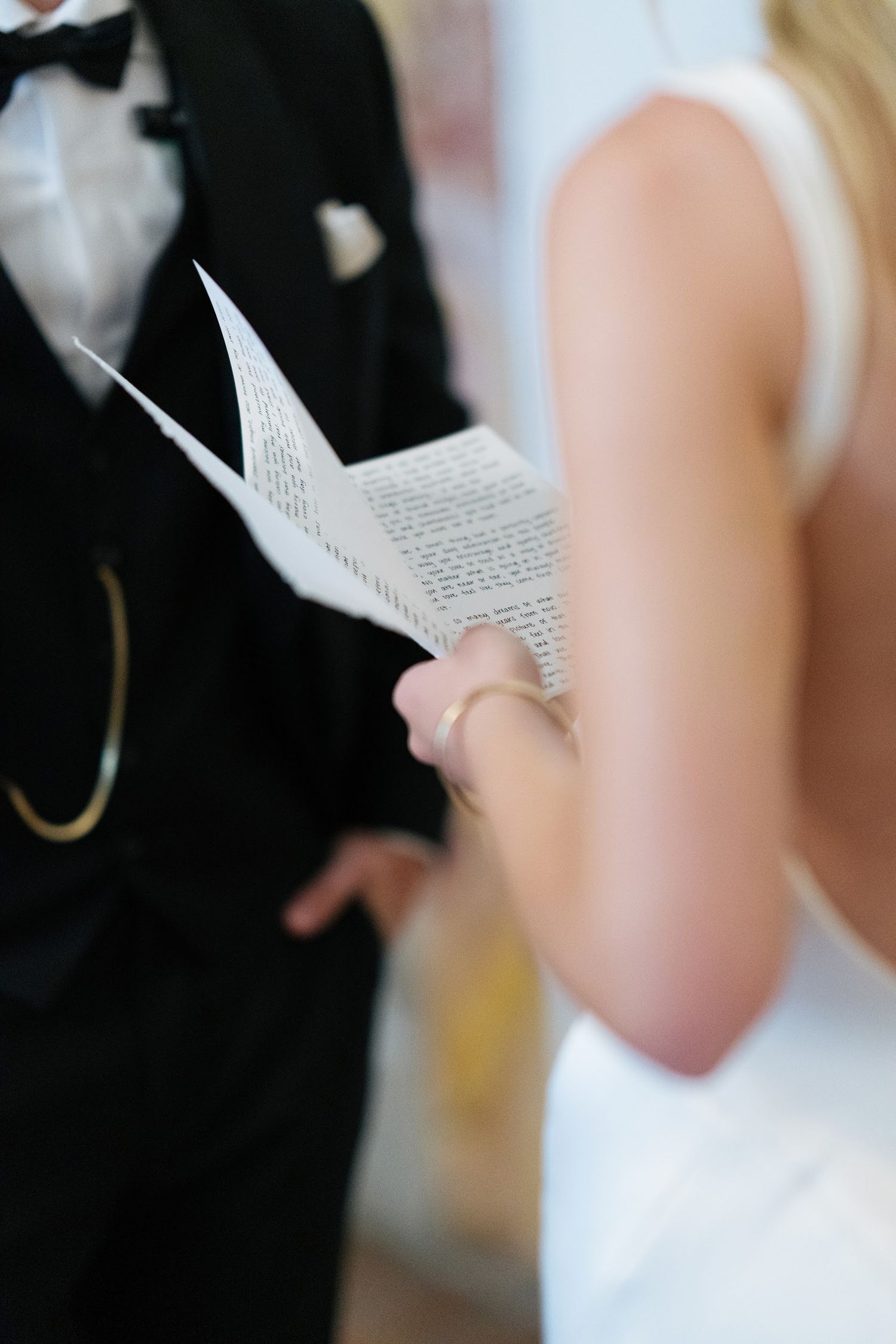 A bride in a white sleeveless gown holds written wedding vows on folded paper during the ceremony.