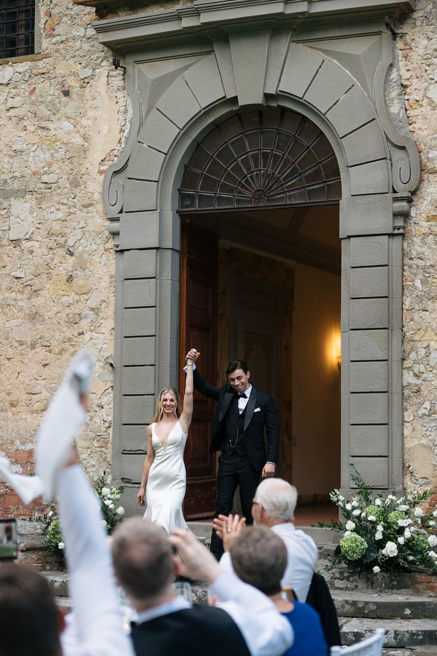Bride and groom celebrate their exit from a classic Italian stone church doorway with cheering guests.