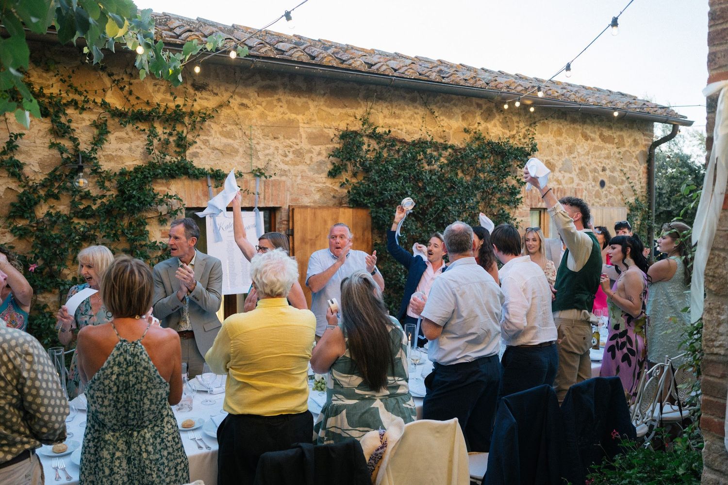 Lively outdoor wedding reception with guests celebrating at long table against rustic stone villa walls.