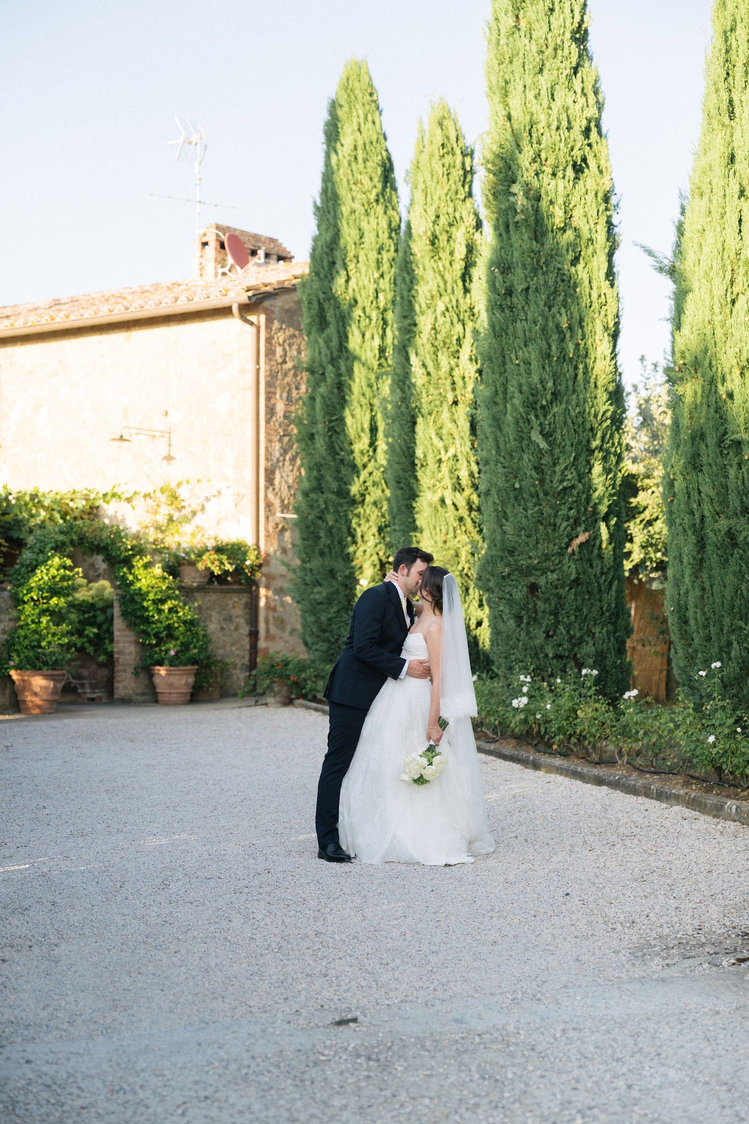 Bride and groom share a kiss near tall cypress trees outside a rustic Tuscan stone villa.