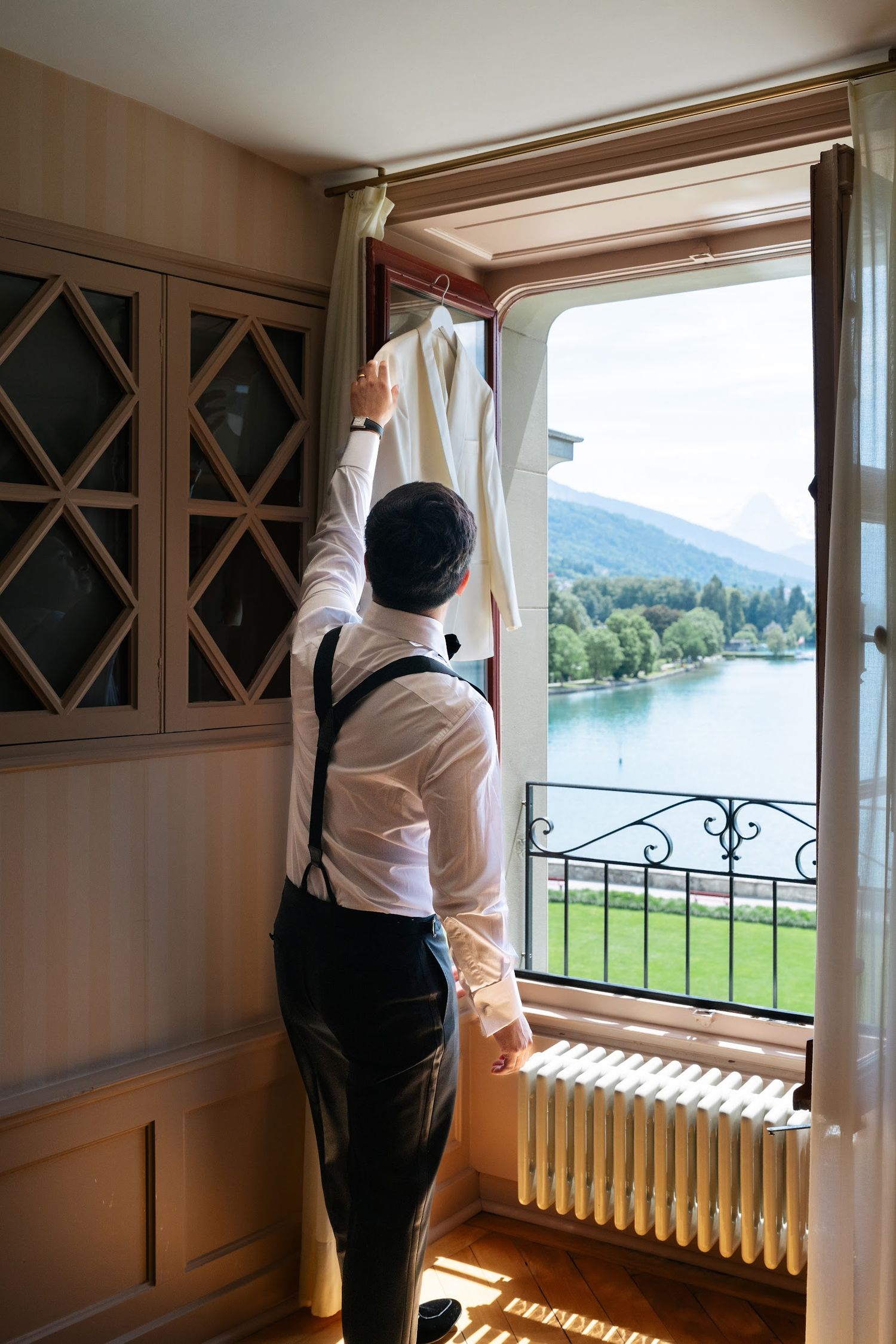 Groom in suspenders hanging his jacket by a window with a scenic lake and mountain view behind him.