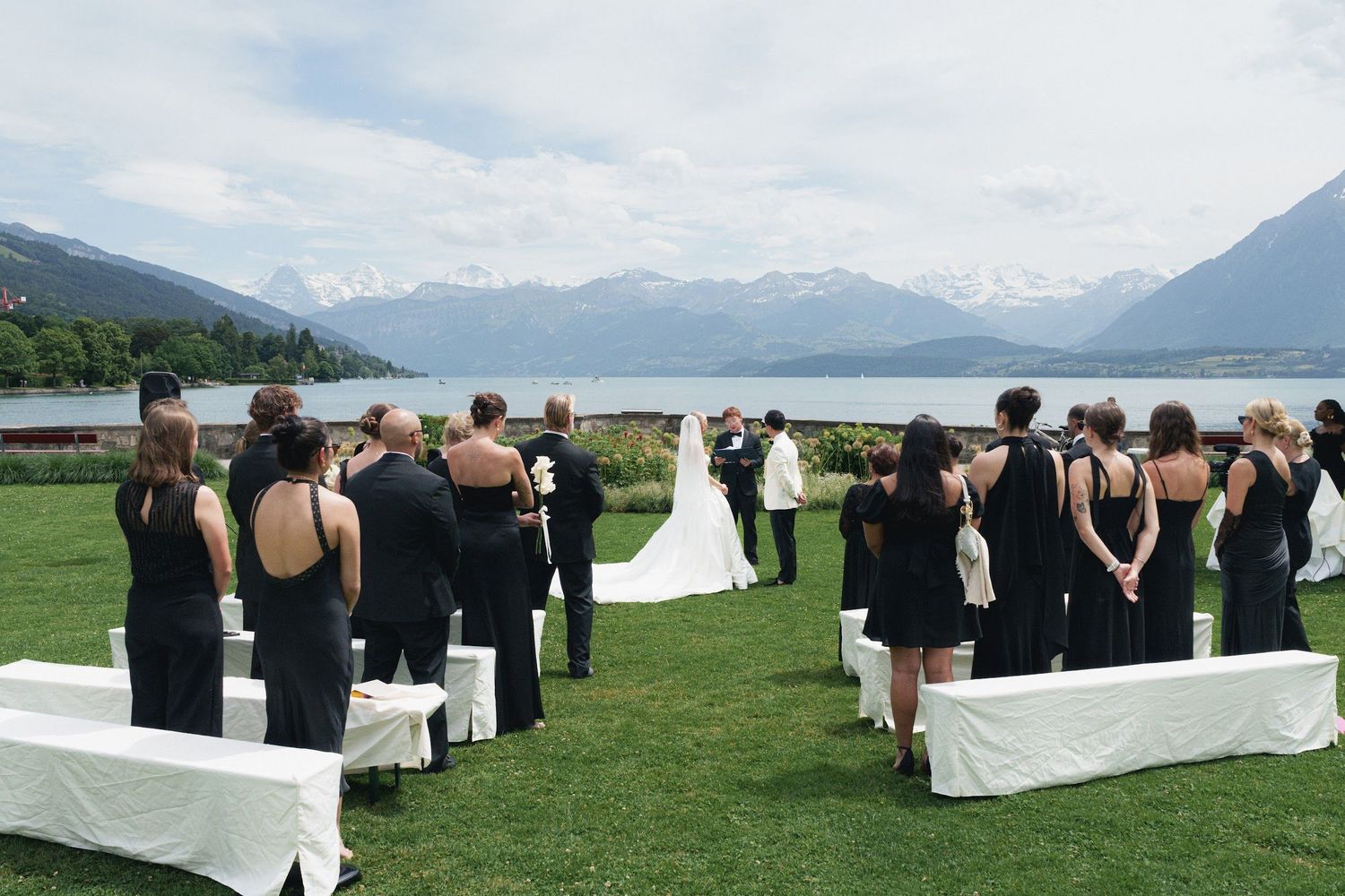 Wedding ceremony on green lawn with guests in black attire, white seating, and stunning alpine lake and mountain views.