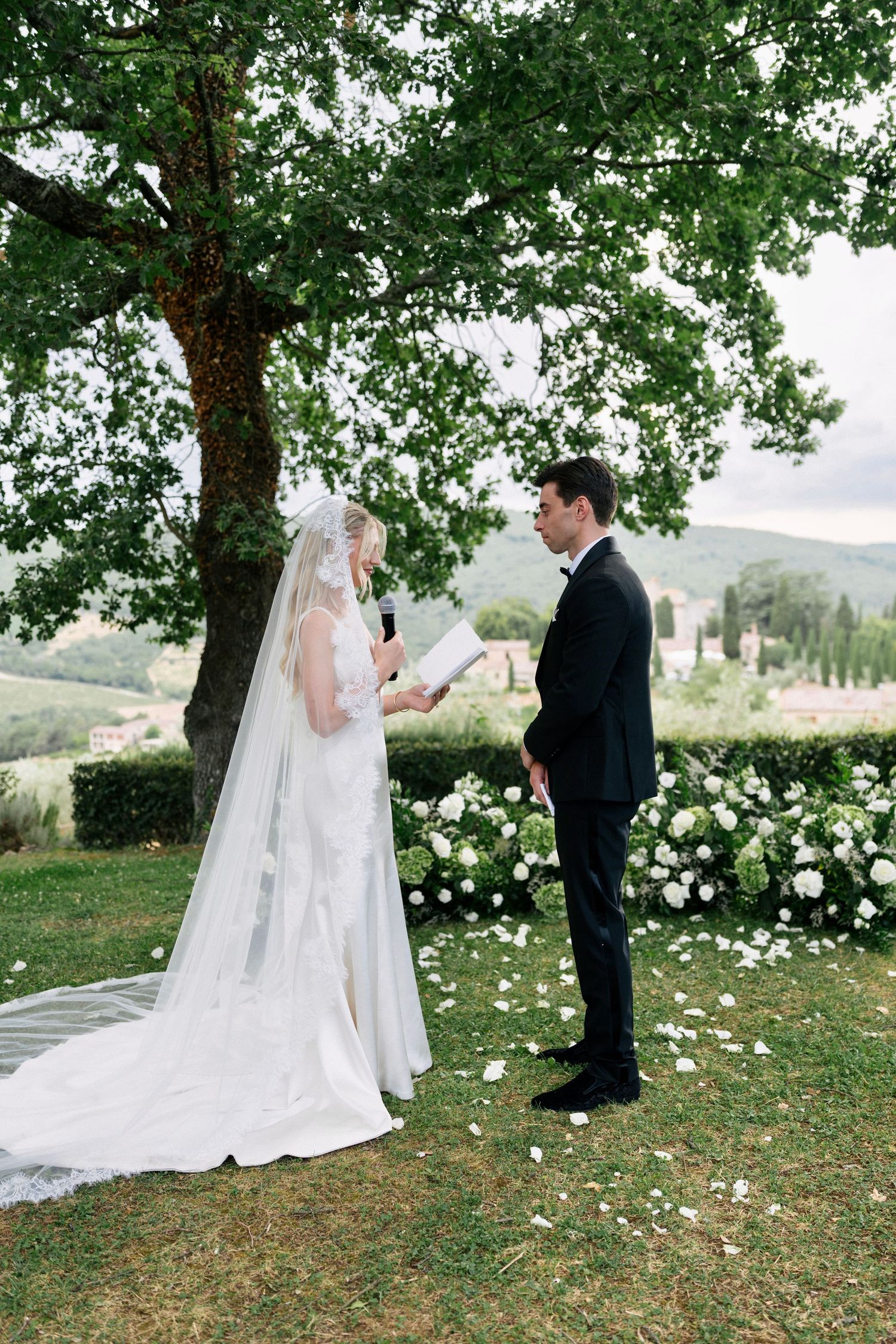 Bride reads vows to groom under large tree with stunning Tuscan countryside and white floral arch backdrop.
