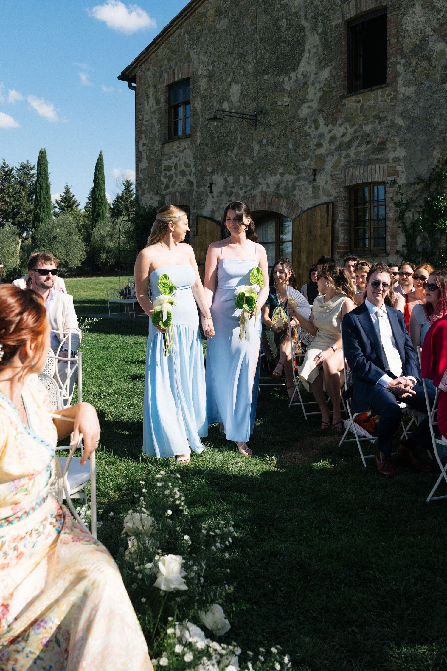 Two bridesmaids in light blue dresses carrying green bouquets walking down outdoor aisle at Tuscan wedding.