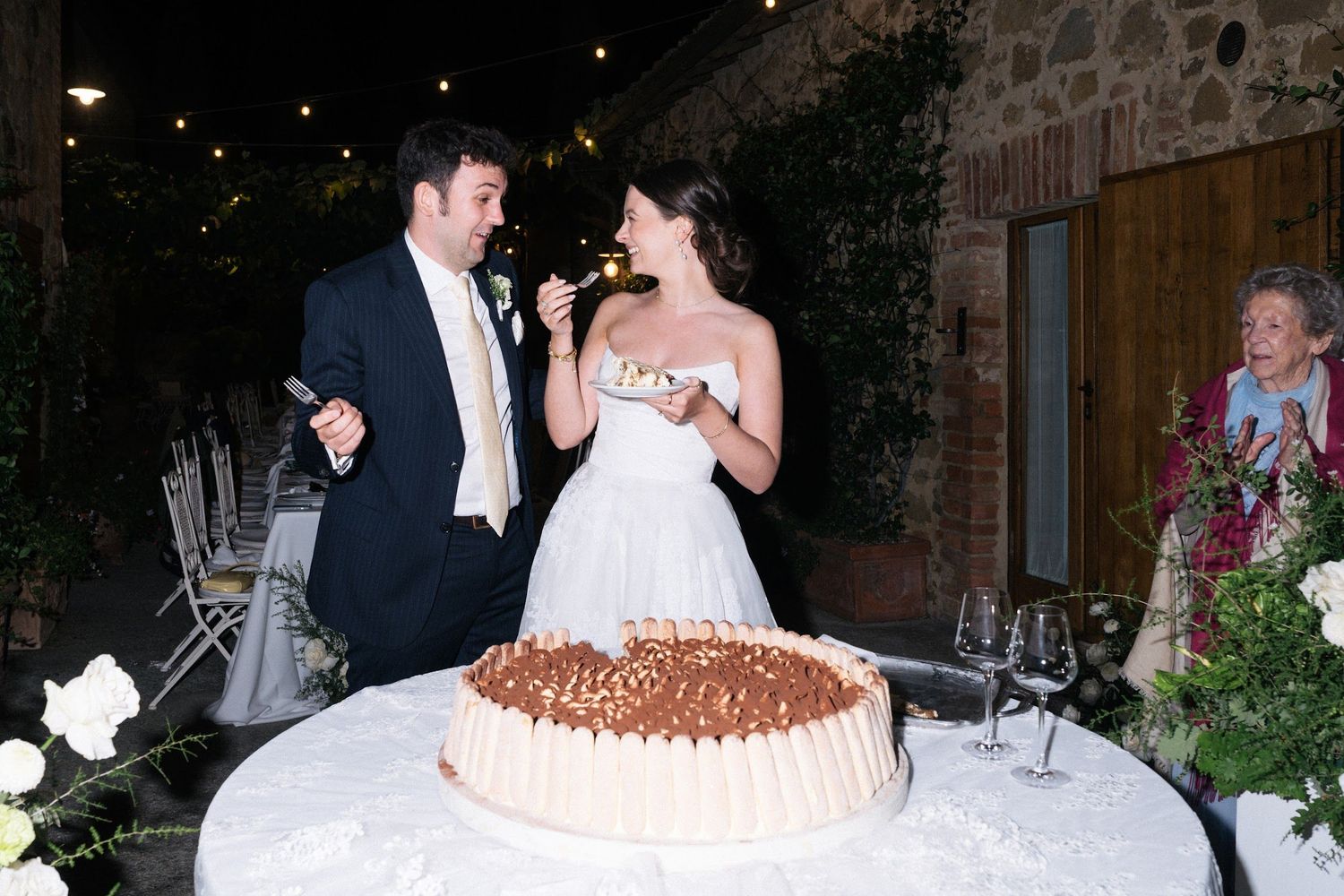 Bride tastes wedding cake as groom watches at outdoor evening reception.