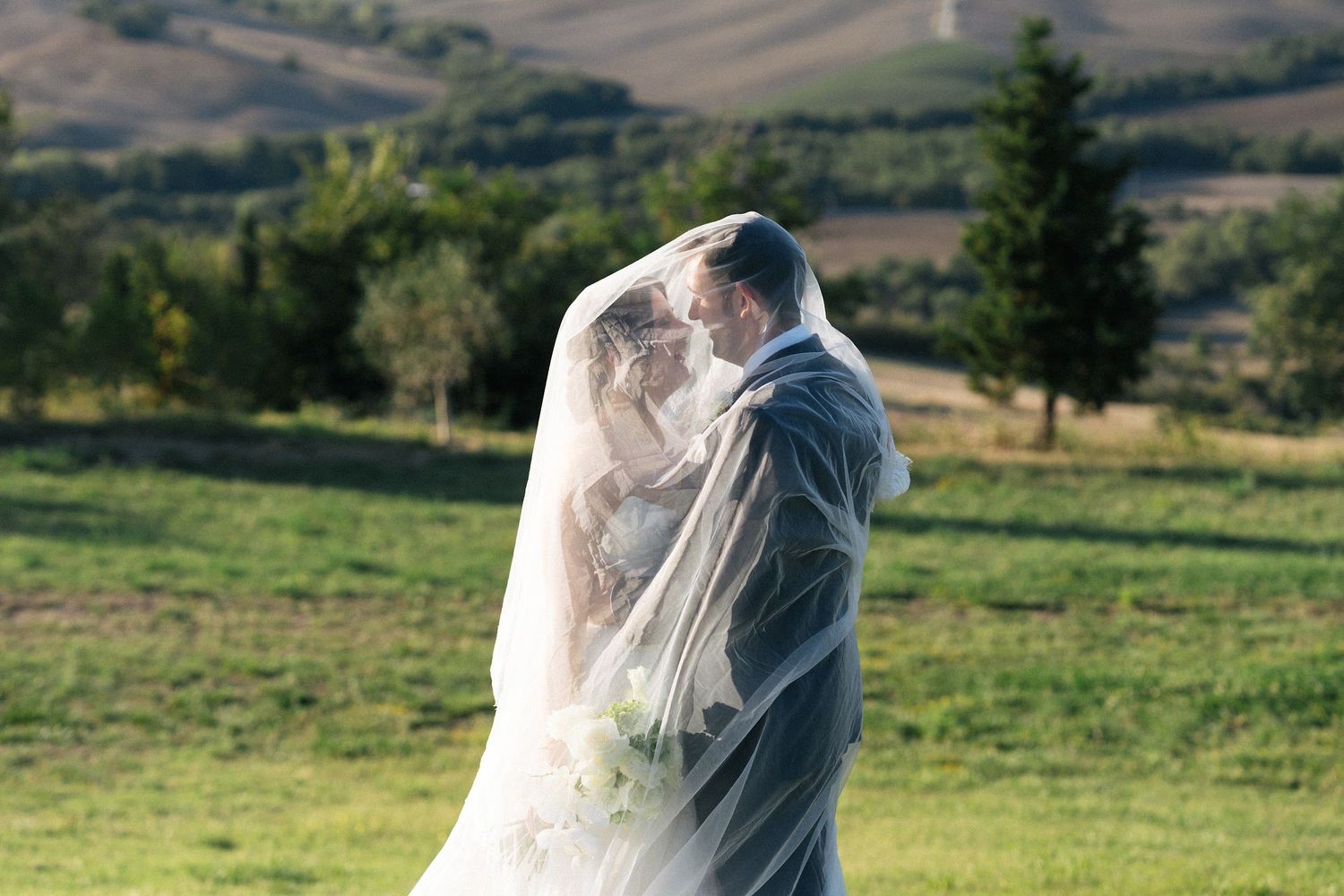 Bride and groom share tender kiss beneath flowing wedding veil in scenic Tuscan countryside setting.