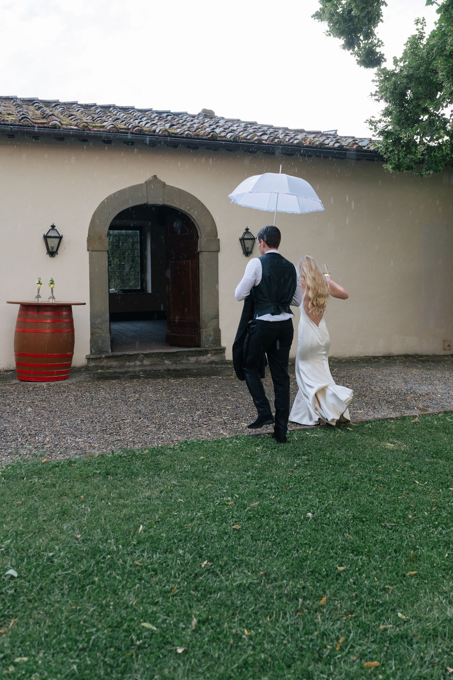 Bride and groom walking together under white umbrella in the rain at outdoor venue.