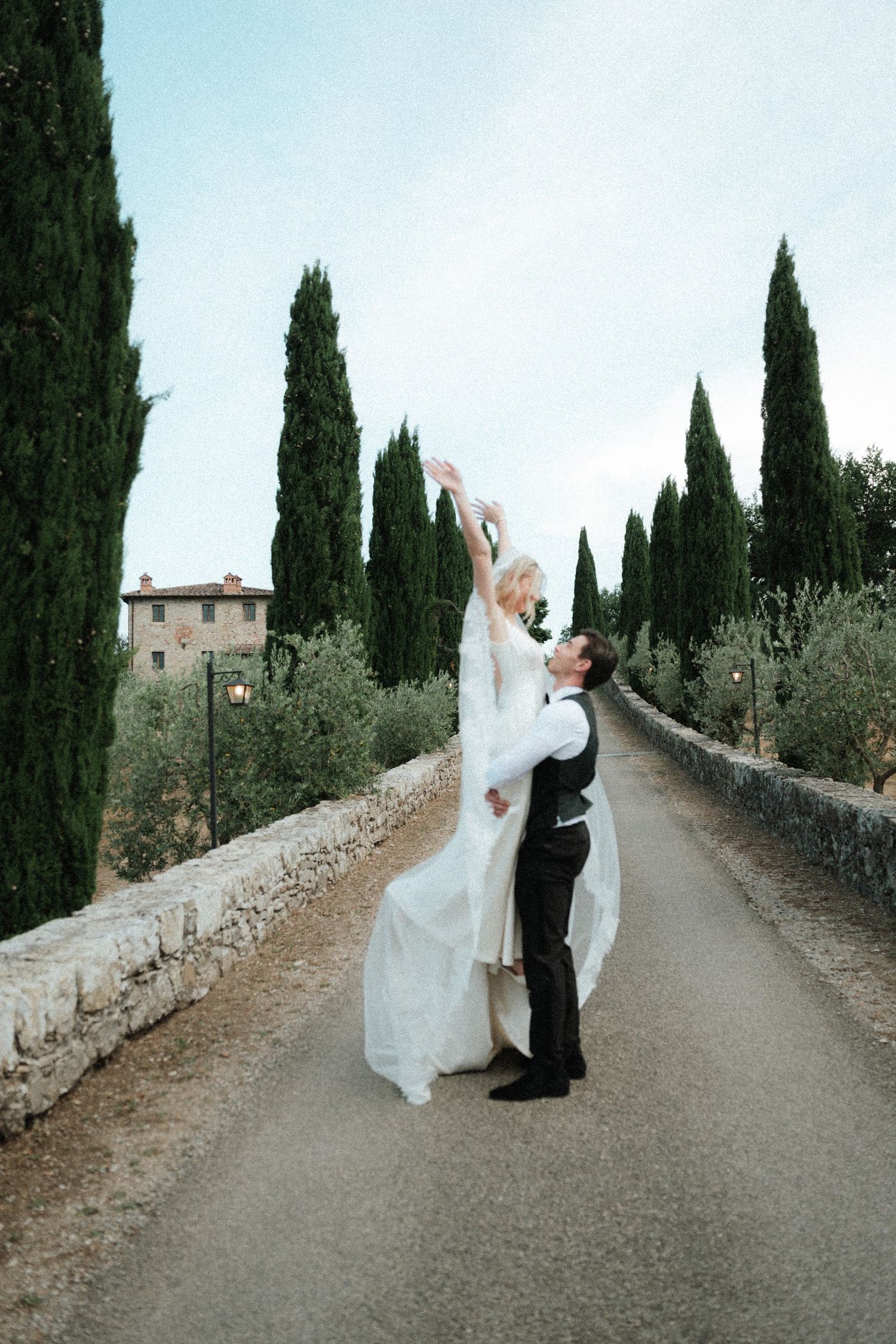 Groom lifts bride in white gown on a cypress-lined road at a romantic Tuscan countryside wedding.