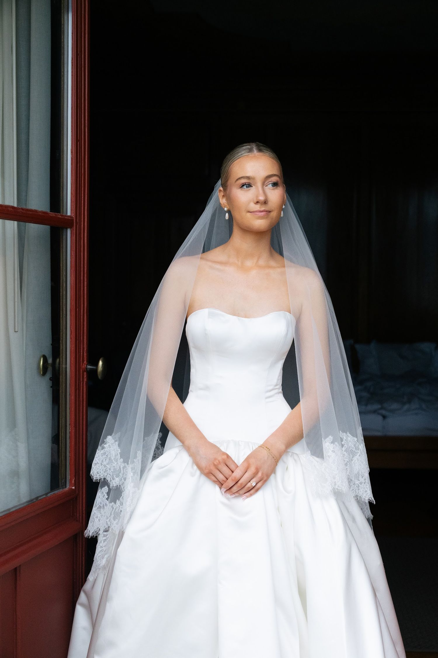 Elegant bride in strapless satin ball gown with lace-edged veil standing in doorway with serene expression.