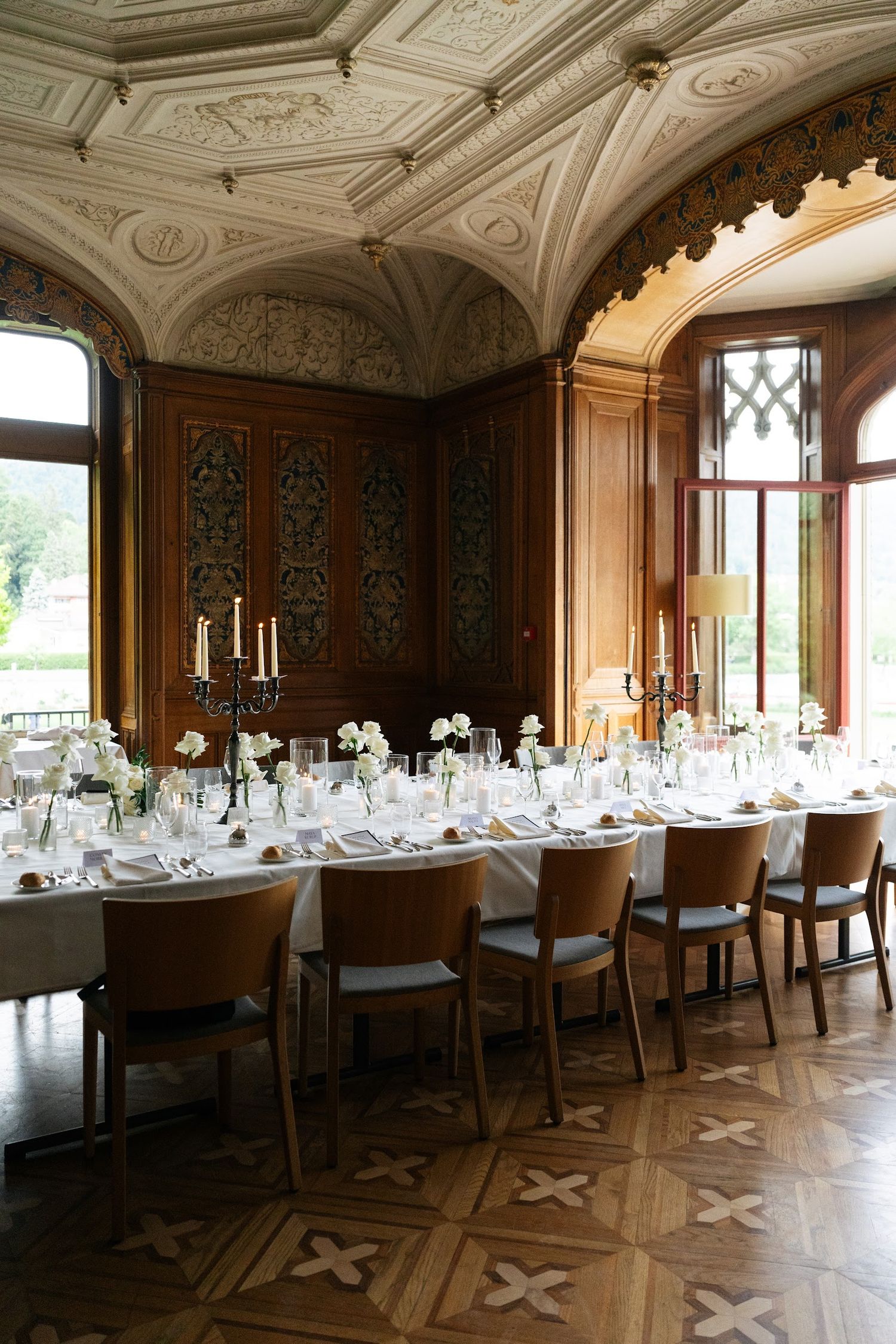 Grand château dining room with ornate plasterwork ceiling, parquet floors, and white floral wedding table setting.