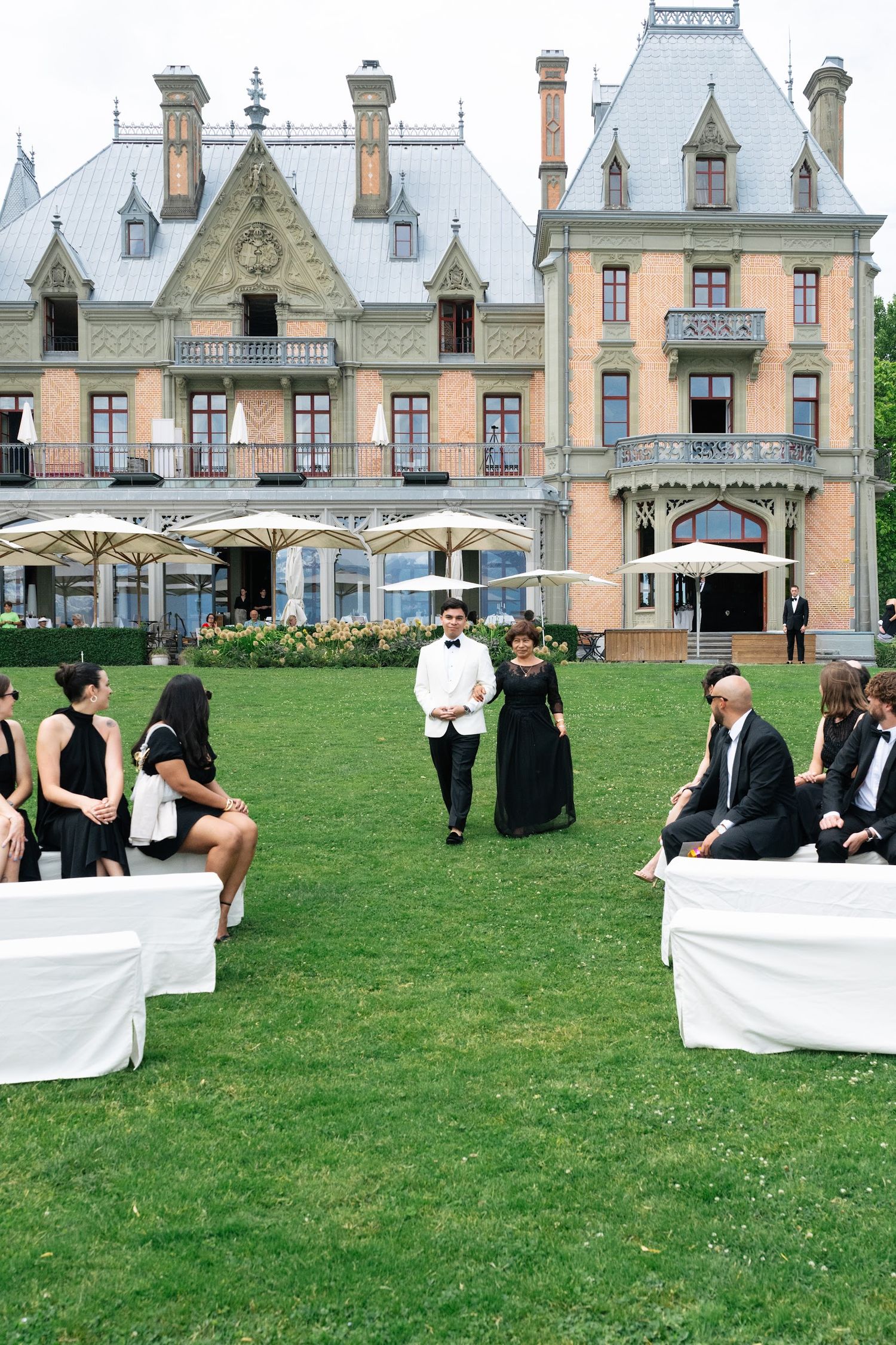 Groom in white tuxedo walks down the aisle between seated guests at an elegant outdoor château wedding ceremony.