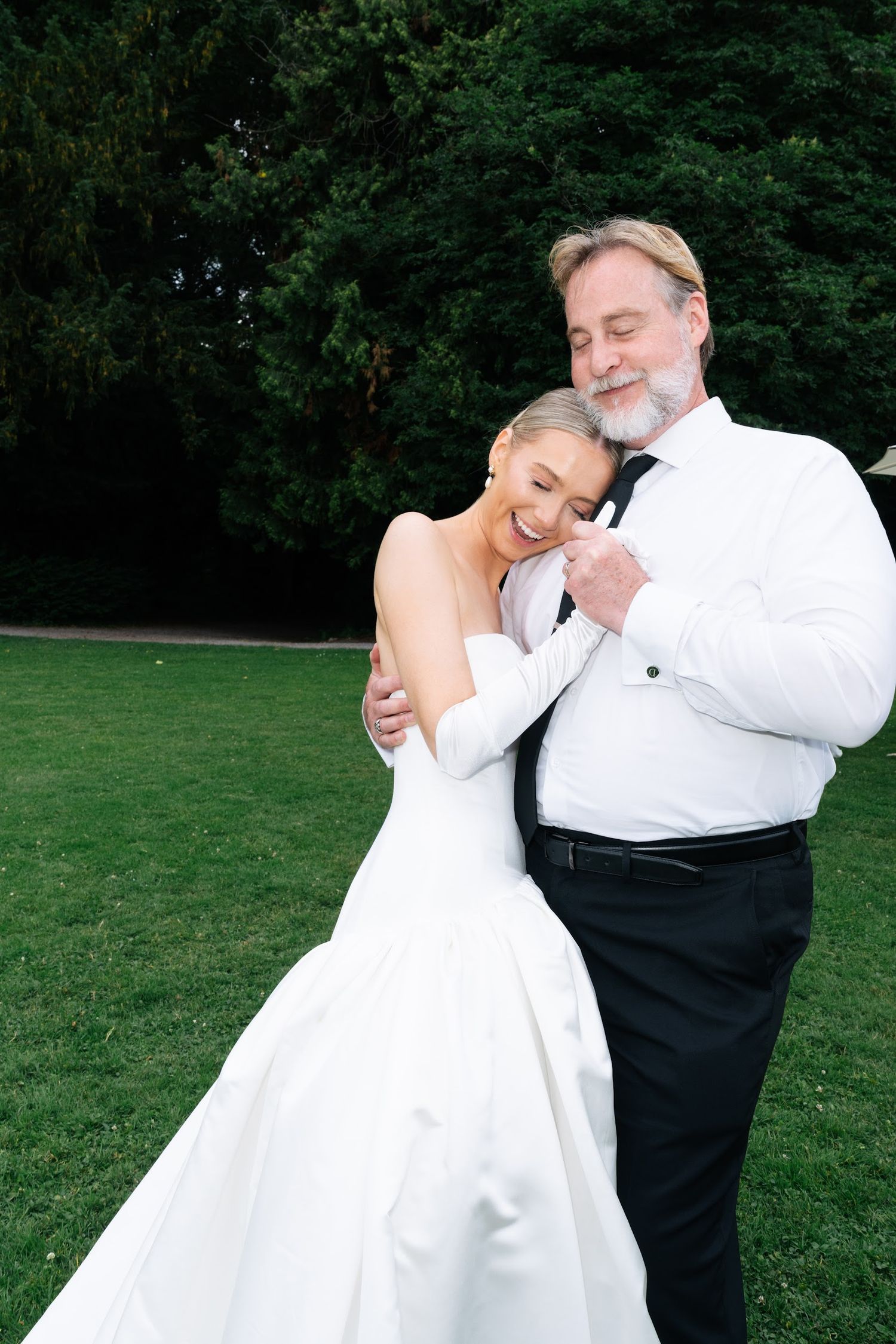 Bride in white strapless gown embracing older man in white shirt and tie on lush green lawn.