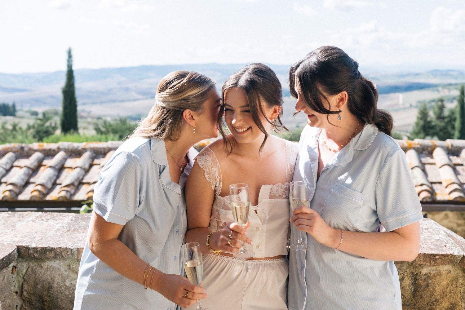 Bride and two bridesmaids in blue robes sharing a joyful embrace while holding champagne glasses outdoors in Tuscany.