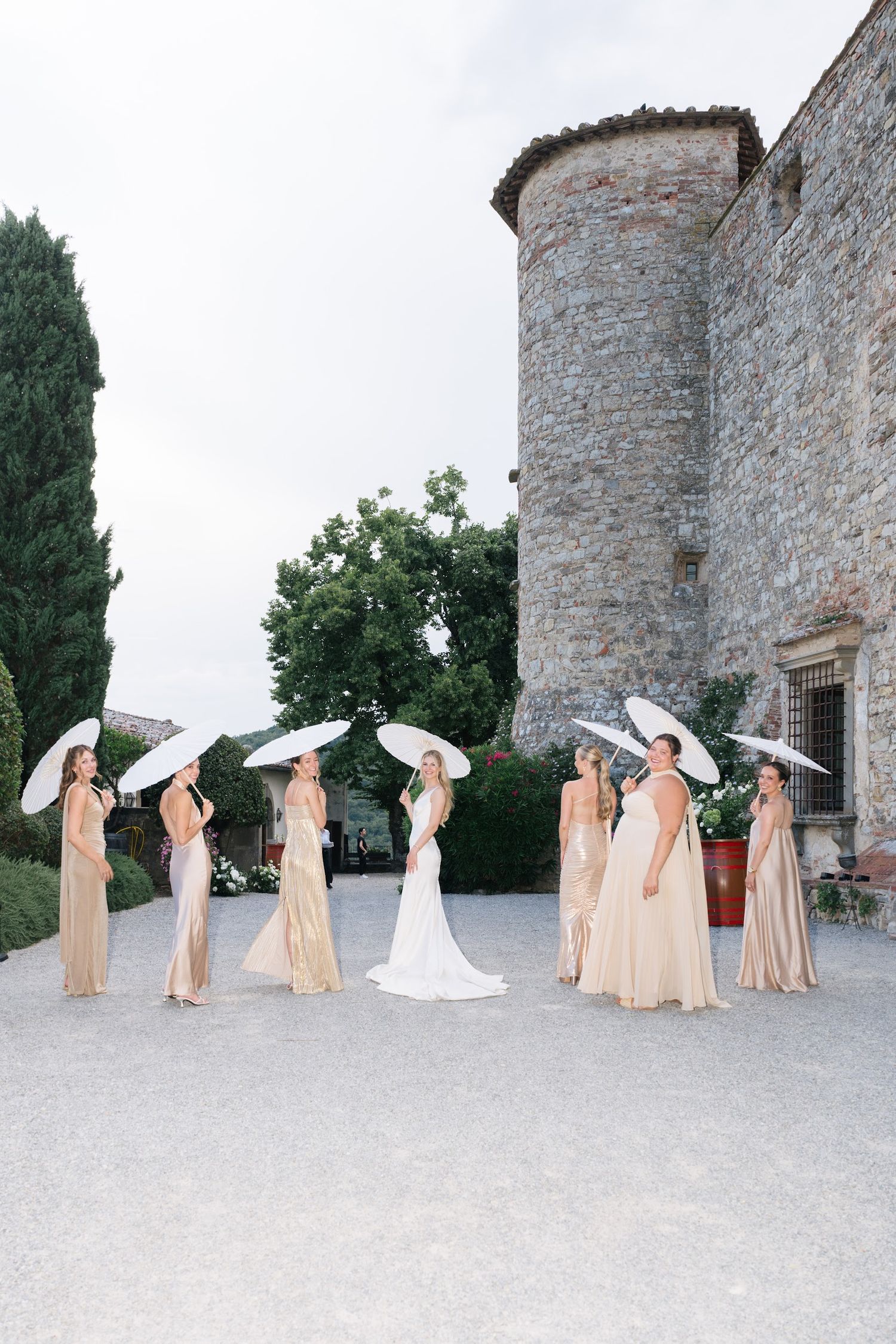 Bride in white gown with bridesmaids in champagne dresses holding white parasols by a stone castle.