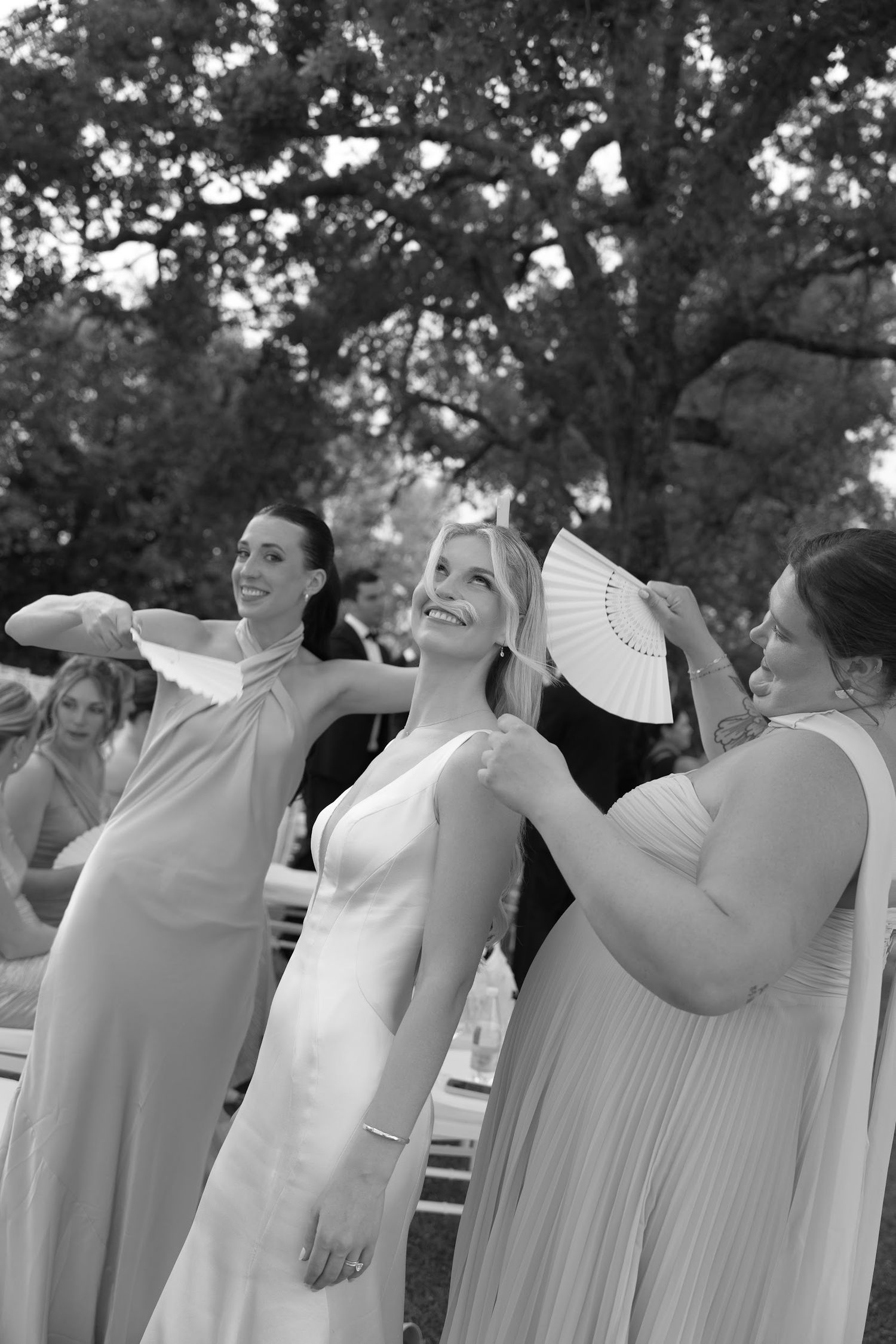 Black and white photo of bride being fanned by bridesmaids in elegant gowns at outdoor wedding.