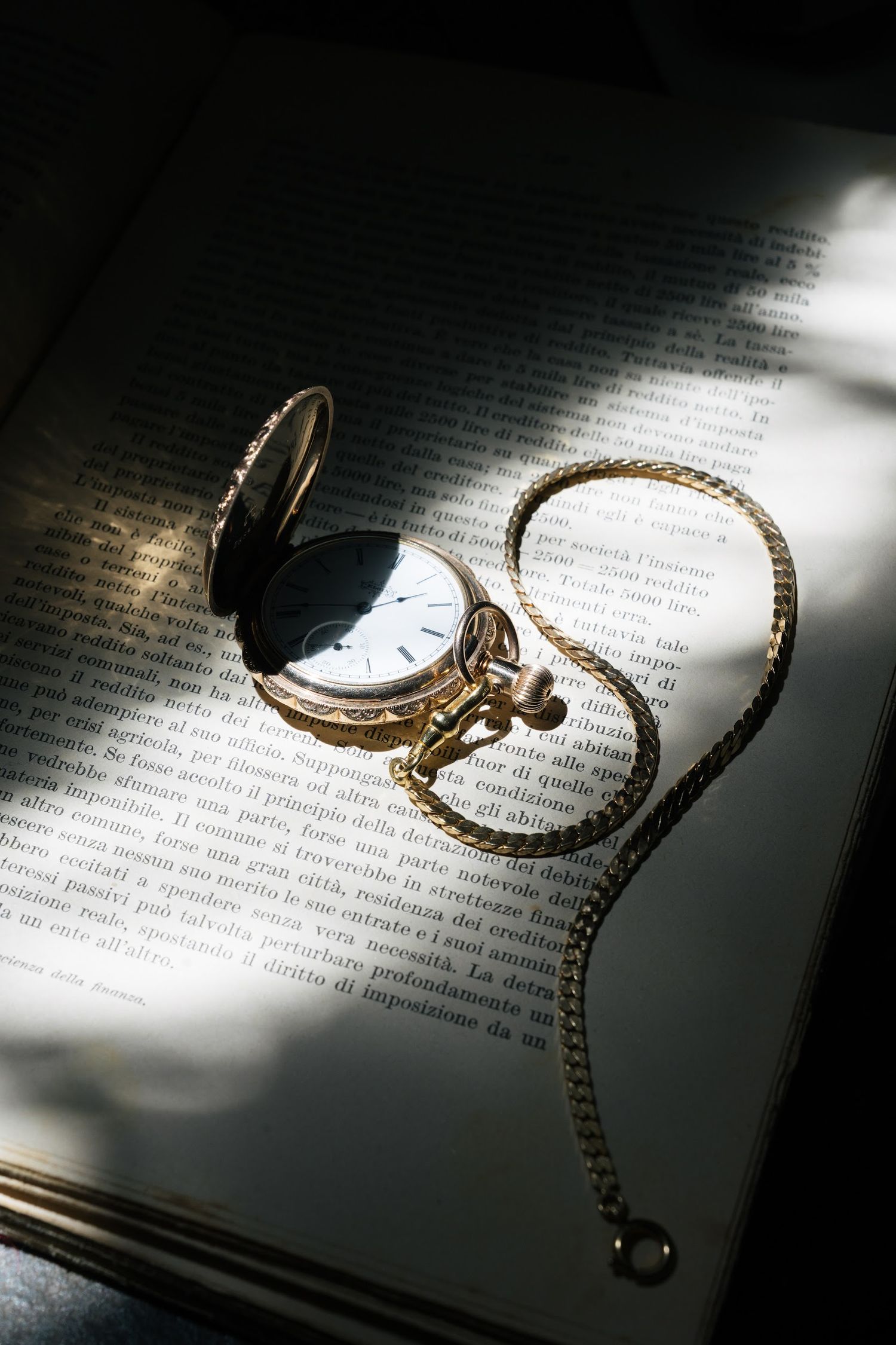 Elegant gold pocket watch with chain resting open on a book page in dramatic natural light.