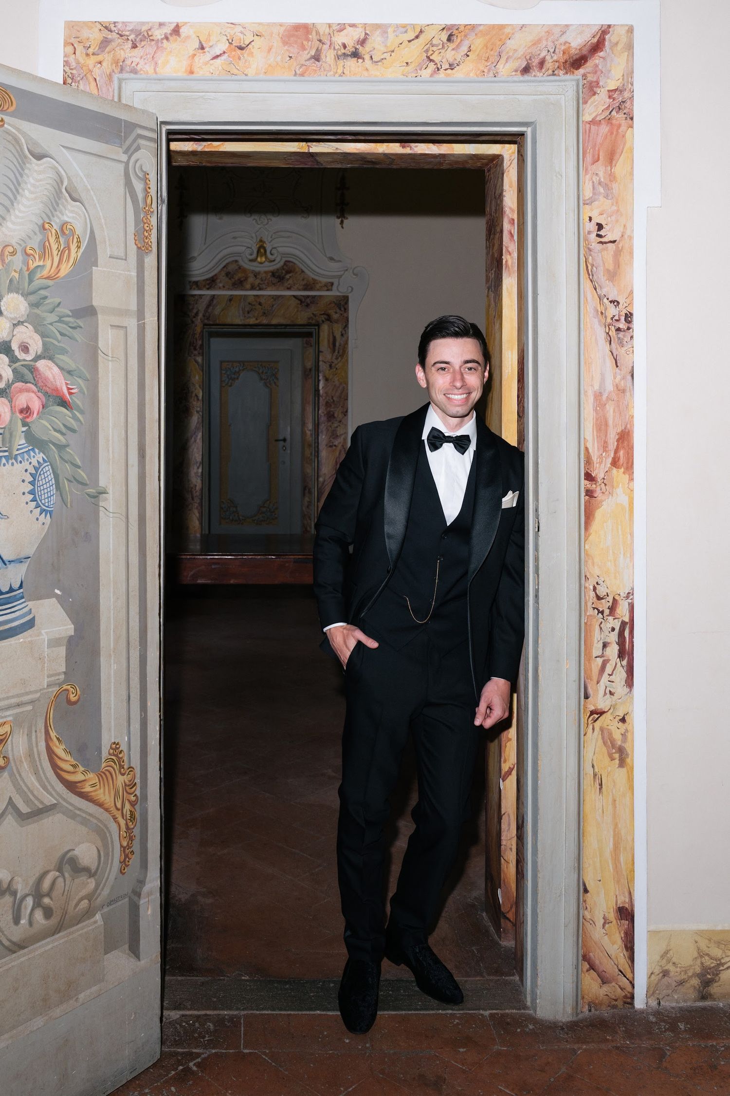 Dapper groom in black tuxedo with bow tie standing in an ornate doorway of a historic Italian villa.