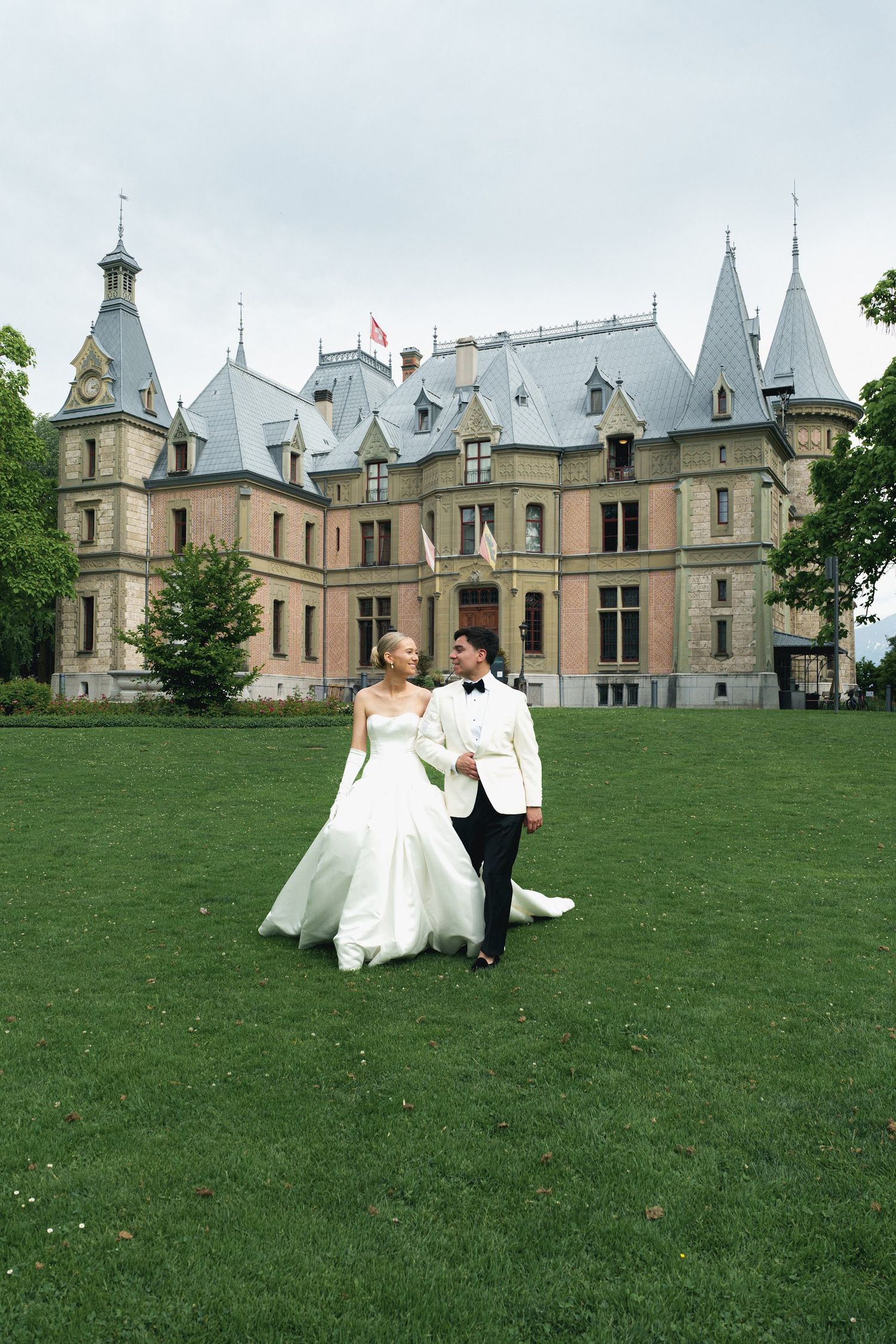 Wedding couple walking hand in hand on lawn before grand château with turrets and slate roof.