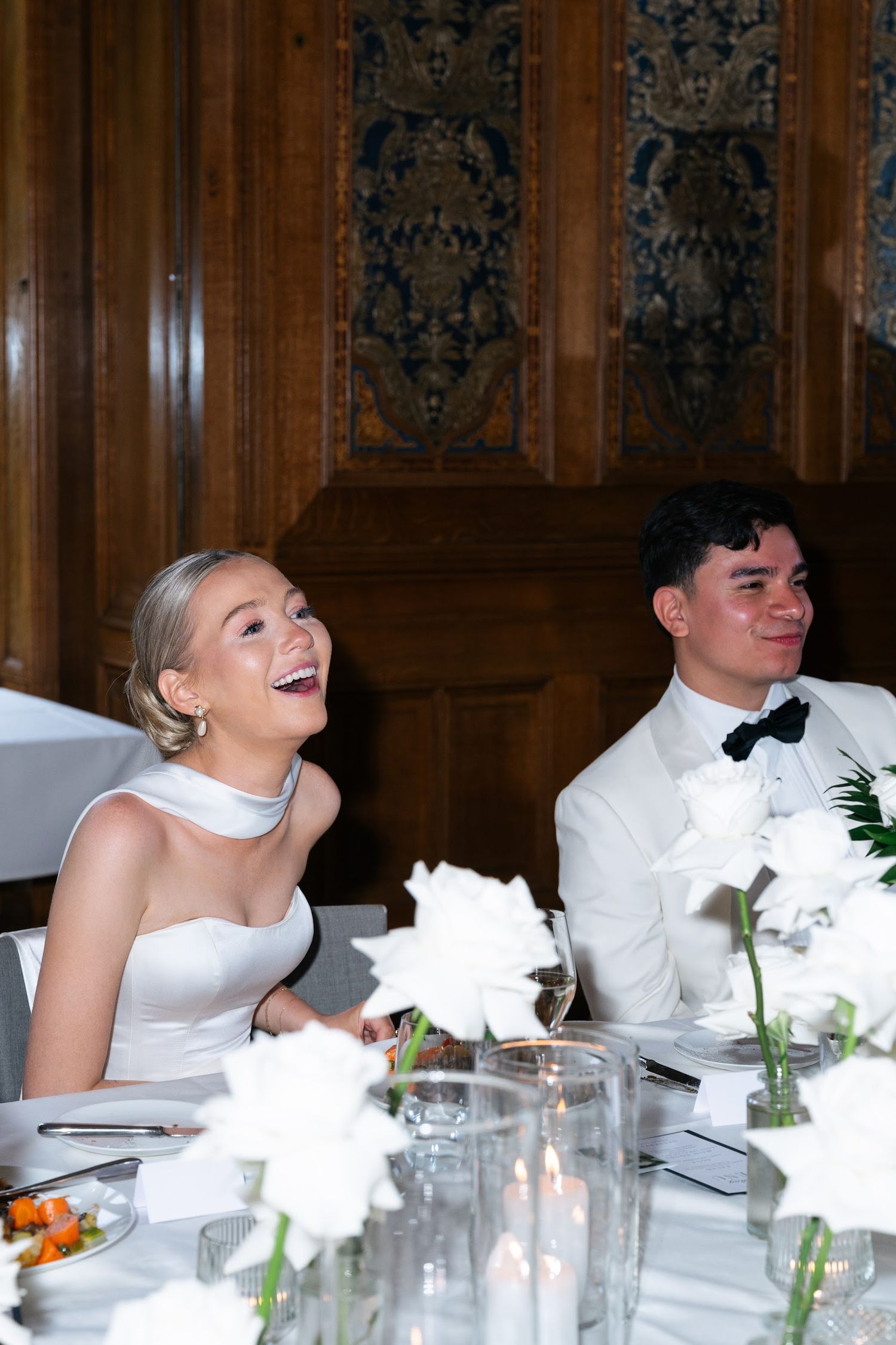 Bride in white strapless gown and groom in white tuxedo laugh joyfully at wedding reception dinner.