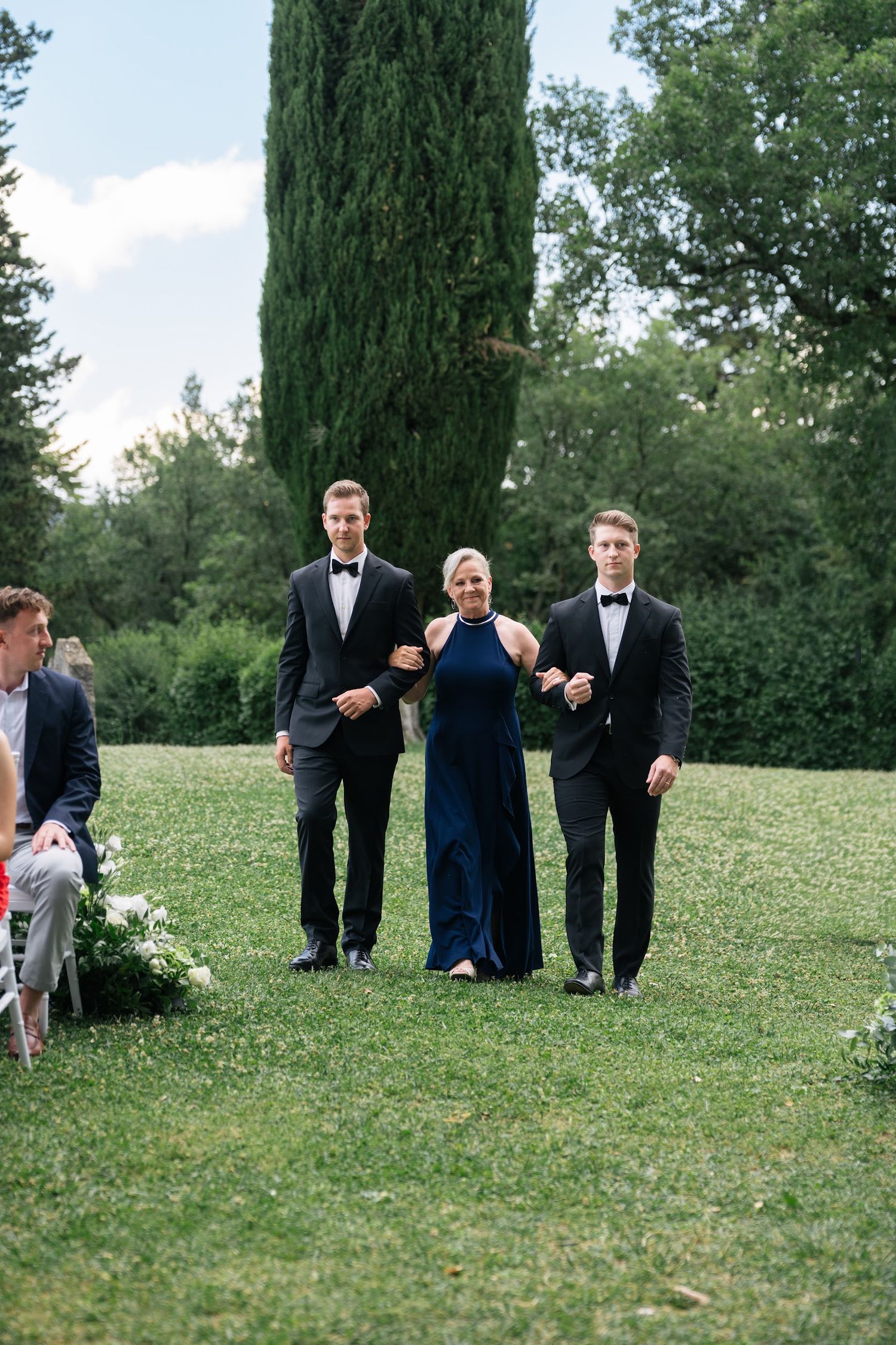 Groomsmen in black tuxedos escort woman in navy gown down aisle at outdoor Italian garden wedding.