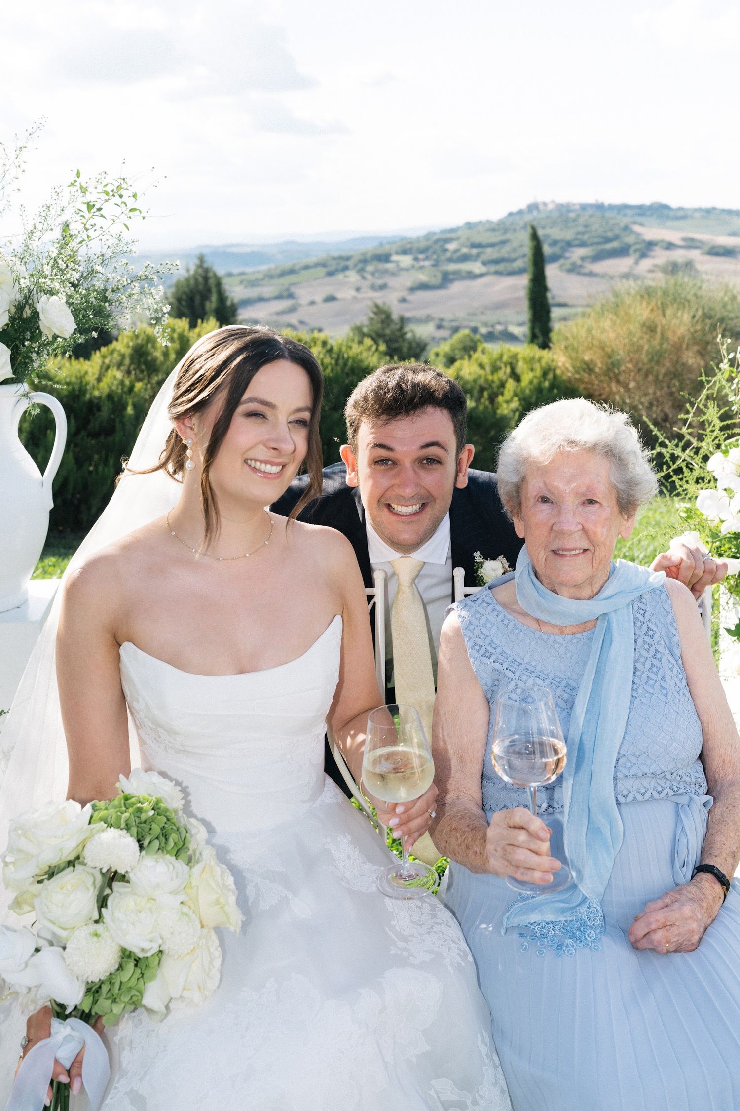 Smiling bride and groom toasting champagne with elderly grandmother at outdoor Tuscan wedding reception.