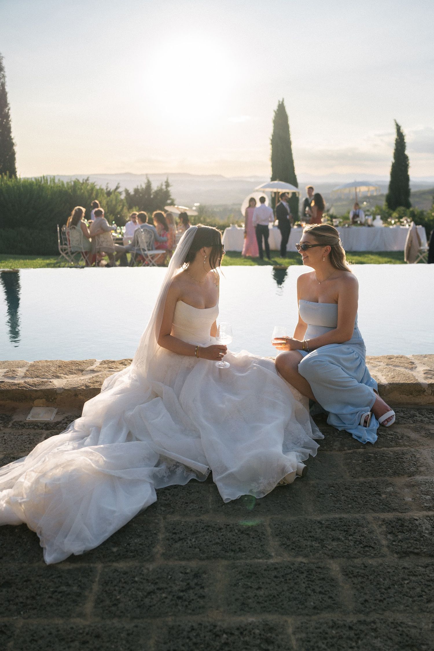 Bride in white gown sits with bridesmaid by infinity pool at golden hour Tuscan villa wedding.