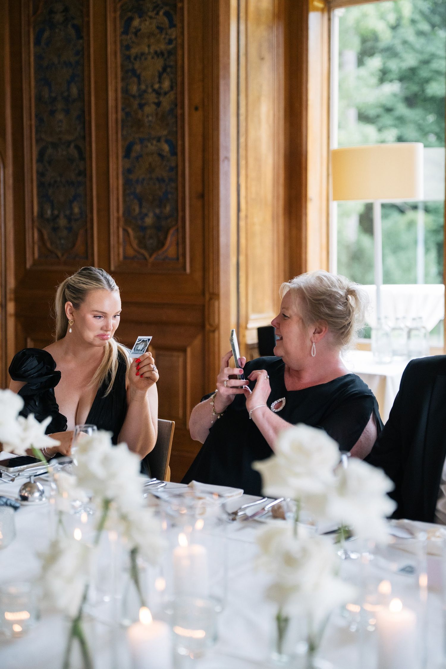 Two women examine photos on phones at a candlelit wedding reception with white floral arrangements.