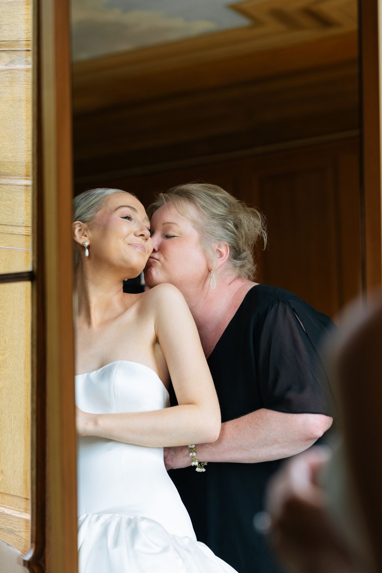Tender moment as mother kisses bride on cheek in doorway before wedding ceremony.