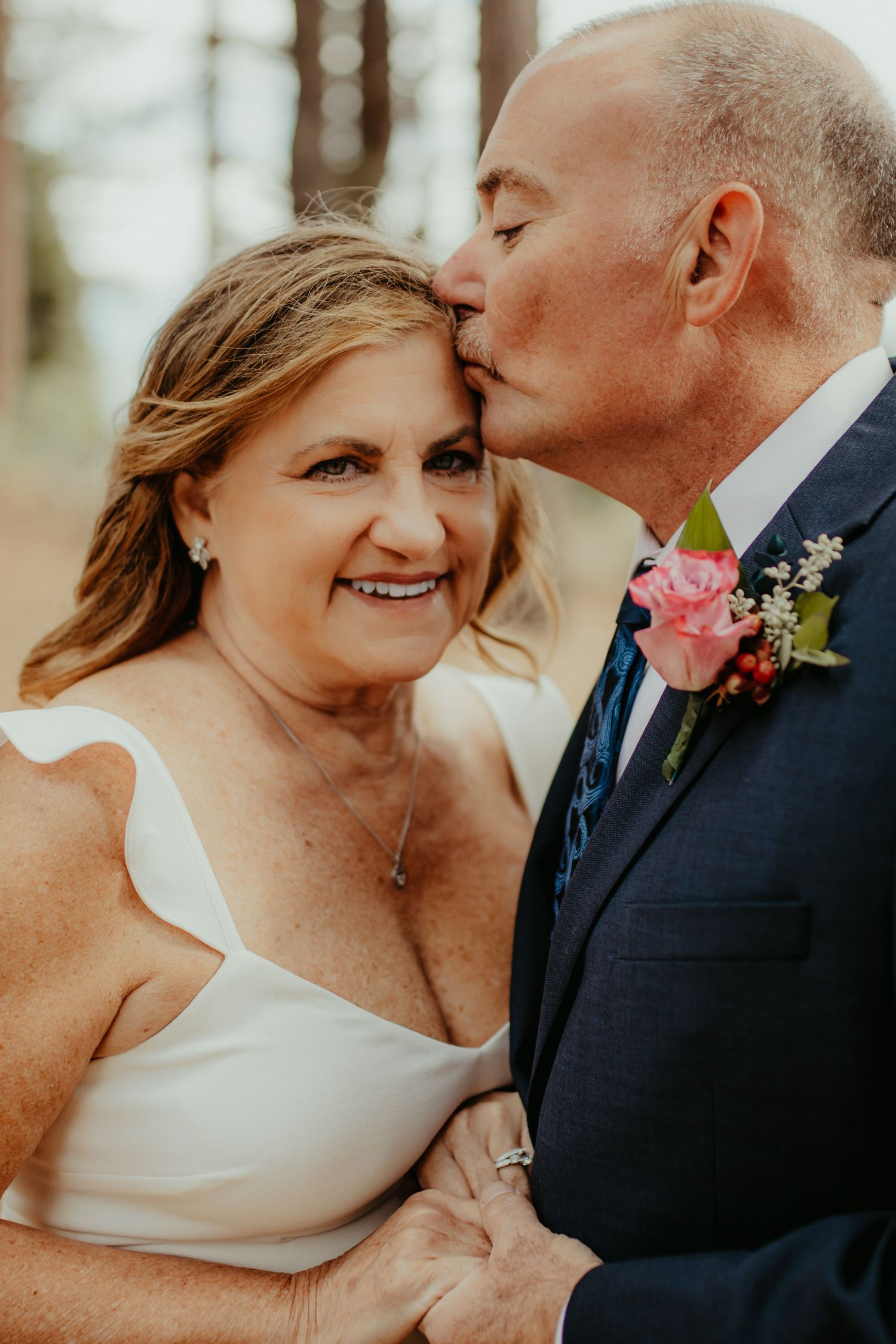 A groom in a navy suit kisses his bride on the forehead as she smiles, both dressed for their wedding day.