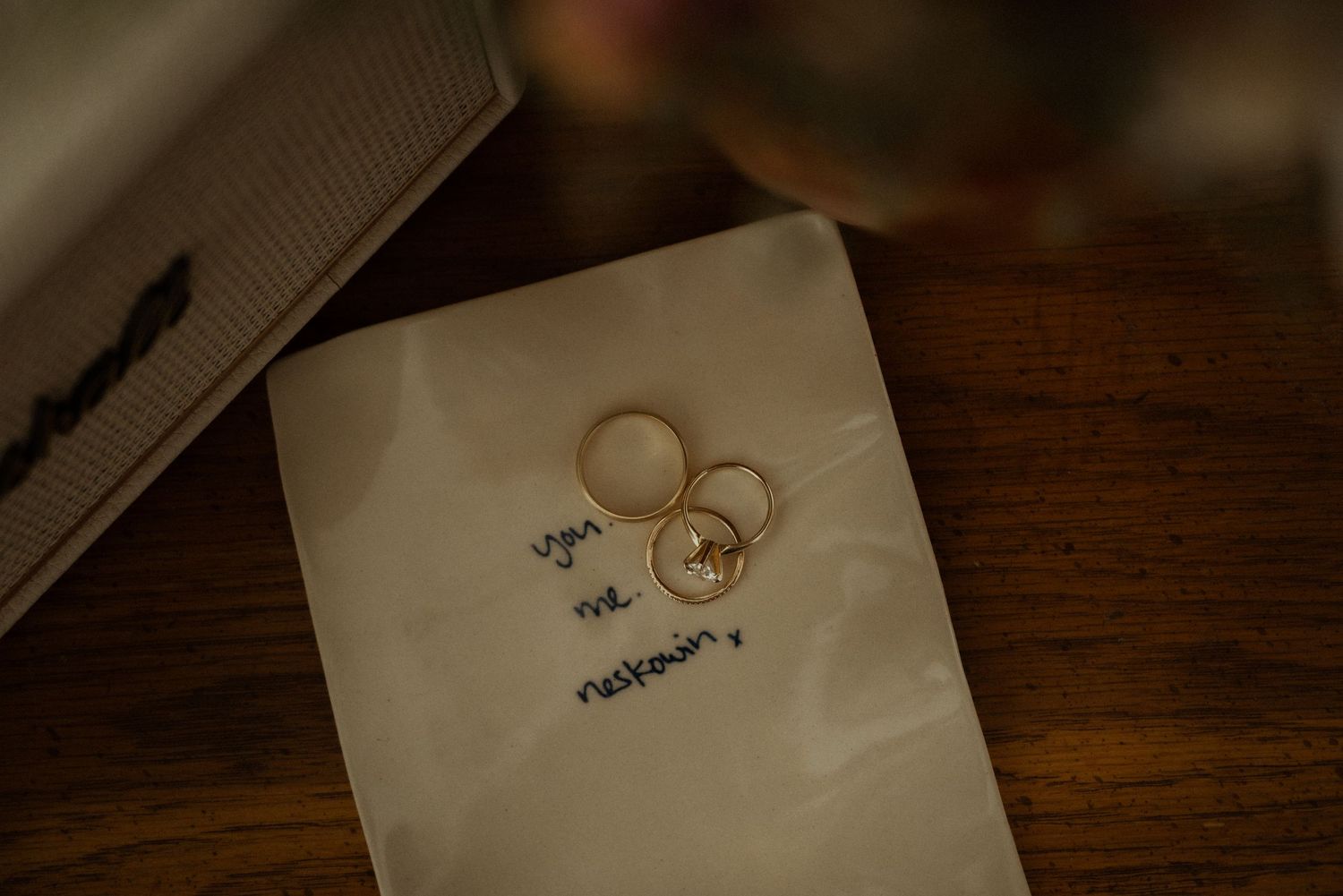 Gold wedding bands and a diamond engagement ring rest on a handwritten note on a rustic wooden table.