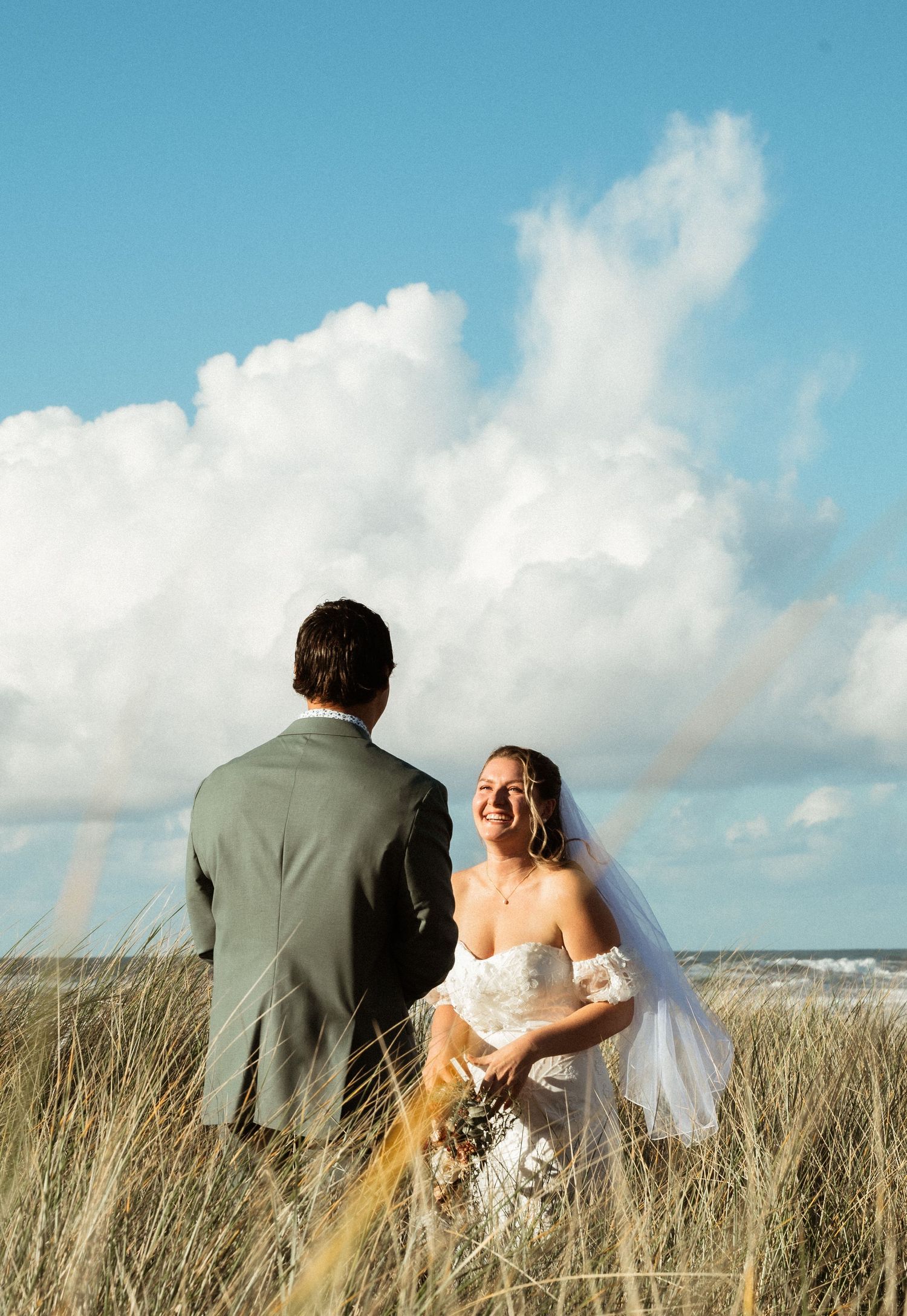 Smiling bride in a white gown and veil faces her groom in a gray suit amid beach grass beneath a bright blue sky.