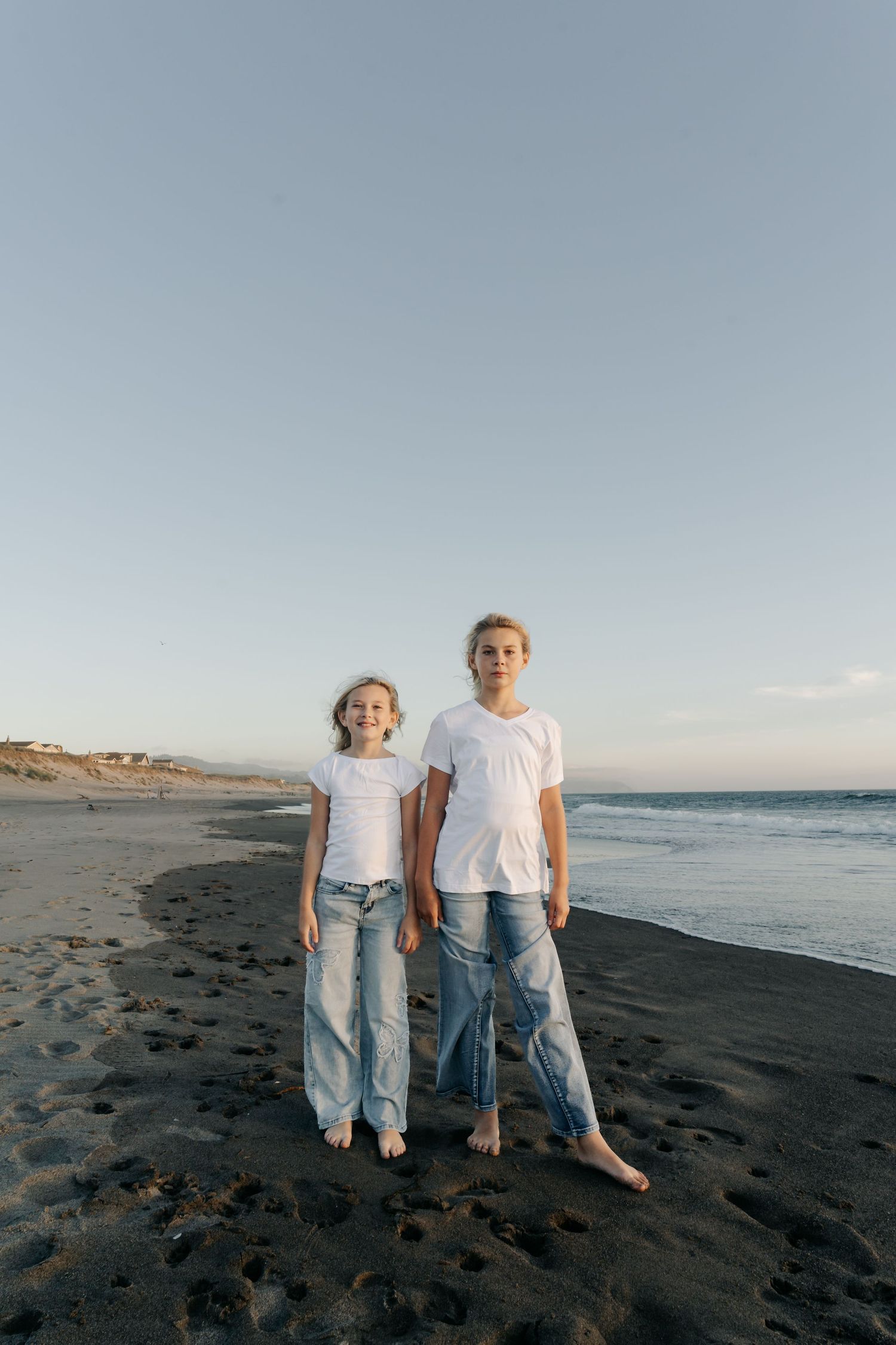 Two children in white t-shirts and jeans standing barefoot on a sandy beach at sunset.