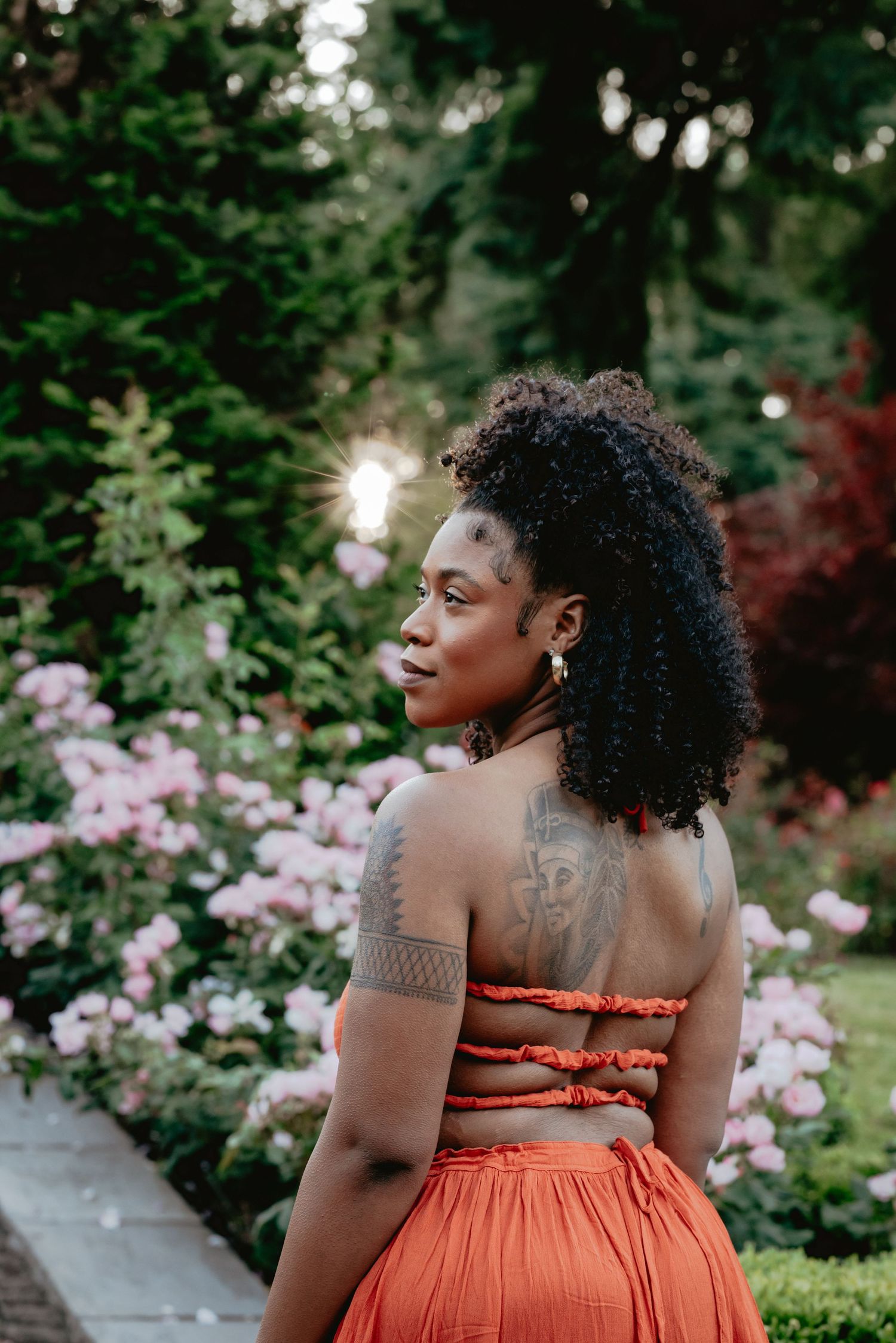 A Black woman in an orange two-piece outfit with back tattoos stands in a lush garden with pink roses.