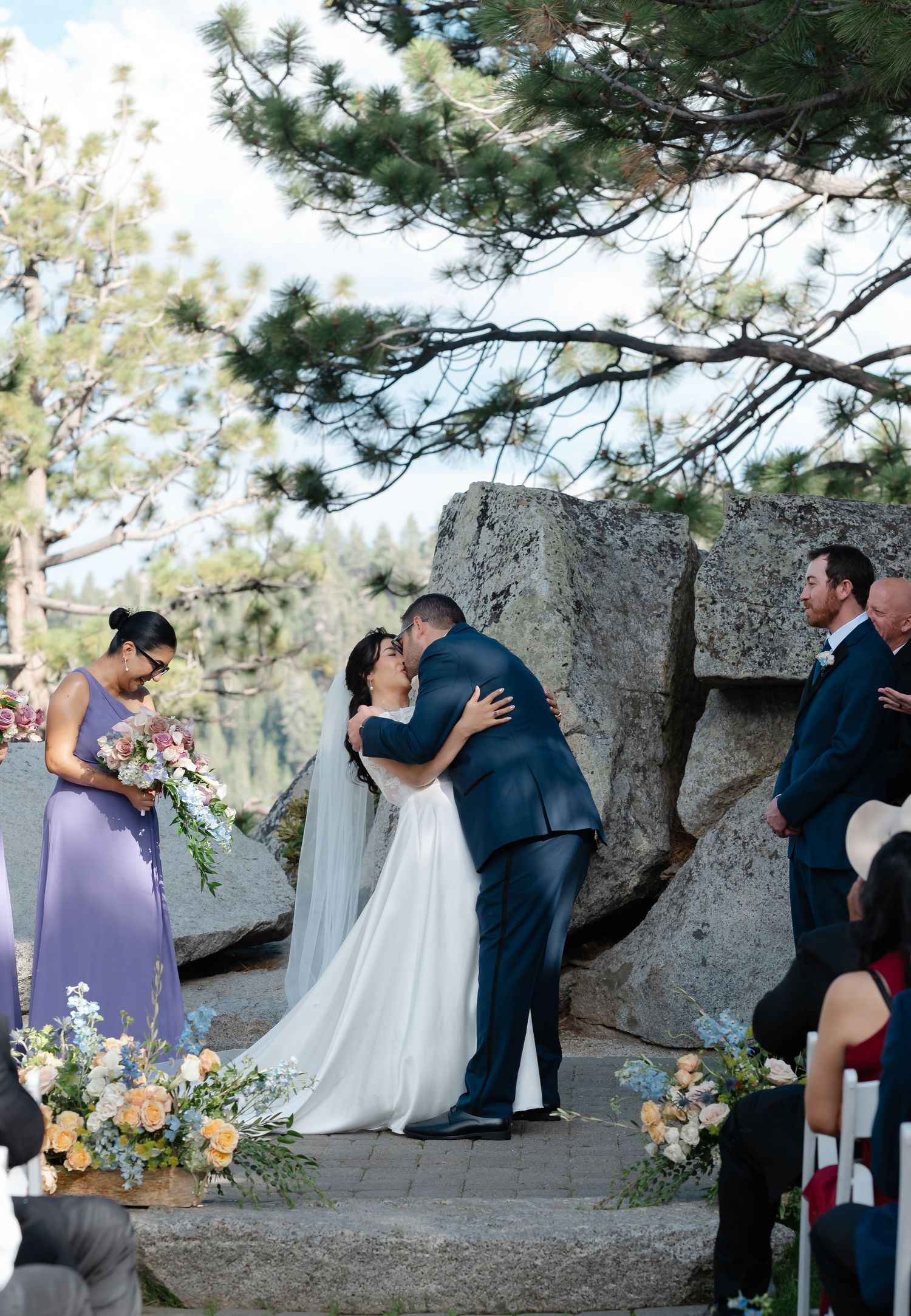 Bride and groom share first kiss at outdoor mountain wedding ceremony with floral arrangements and large granite rocks.