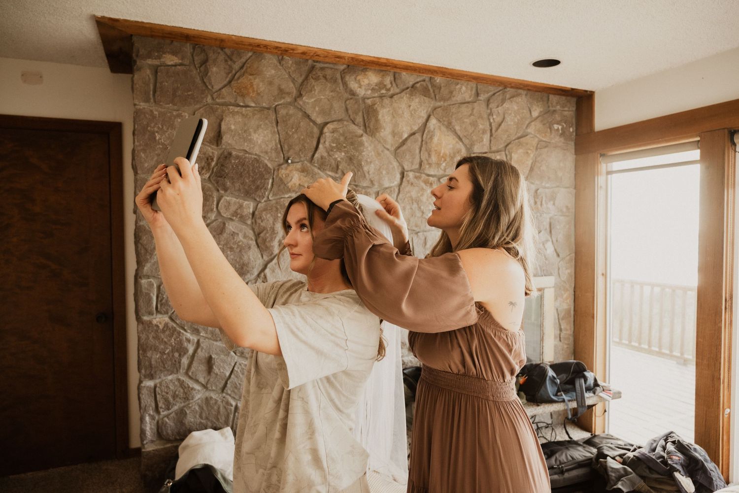 Bridesmaid in a mauve dress styles a woman's hair as she takes a selfie with her phone, in a cozy room with a stone wall.